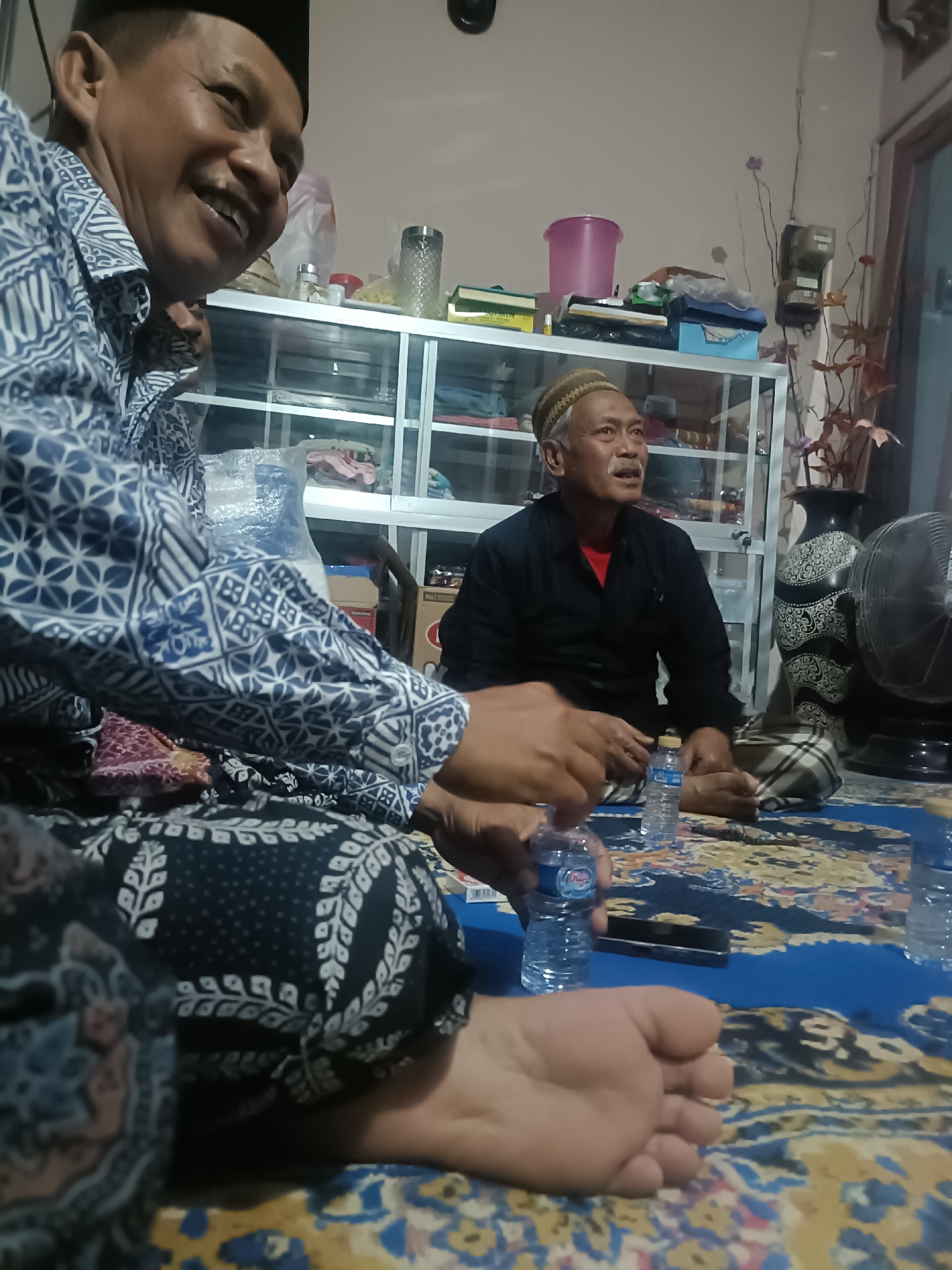 The image depicts an indoor scene, likely a residence in Kediri City, Indonesia. Two adult males are seated on the floor on a blue and yellow patterned carpet. The man on the left is in the foreground, partially out of frame, wearing a blue and white patterned shirt and a black cap. He is smiling and looking to his right. The man on the right is in the middle ground, wearing a black shirt and a woven cap. He has a mustache and appears to be speaking or looking towards something off-camera. Several plastic water bottles are visible on the carpet between the two men, one being held by the man on the left. In the background, there is a white glass-fronted cabinet filled with various items, including what appears to be clothing or textiles. A black fan is positioned to the right of the man in the black shirt, and a decorative vase with intricate patterns is next to it. The lighting suggests it is evening or nighttime.