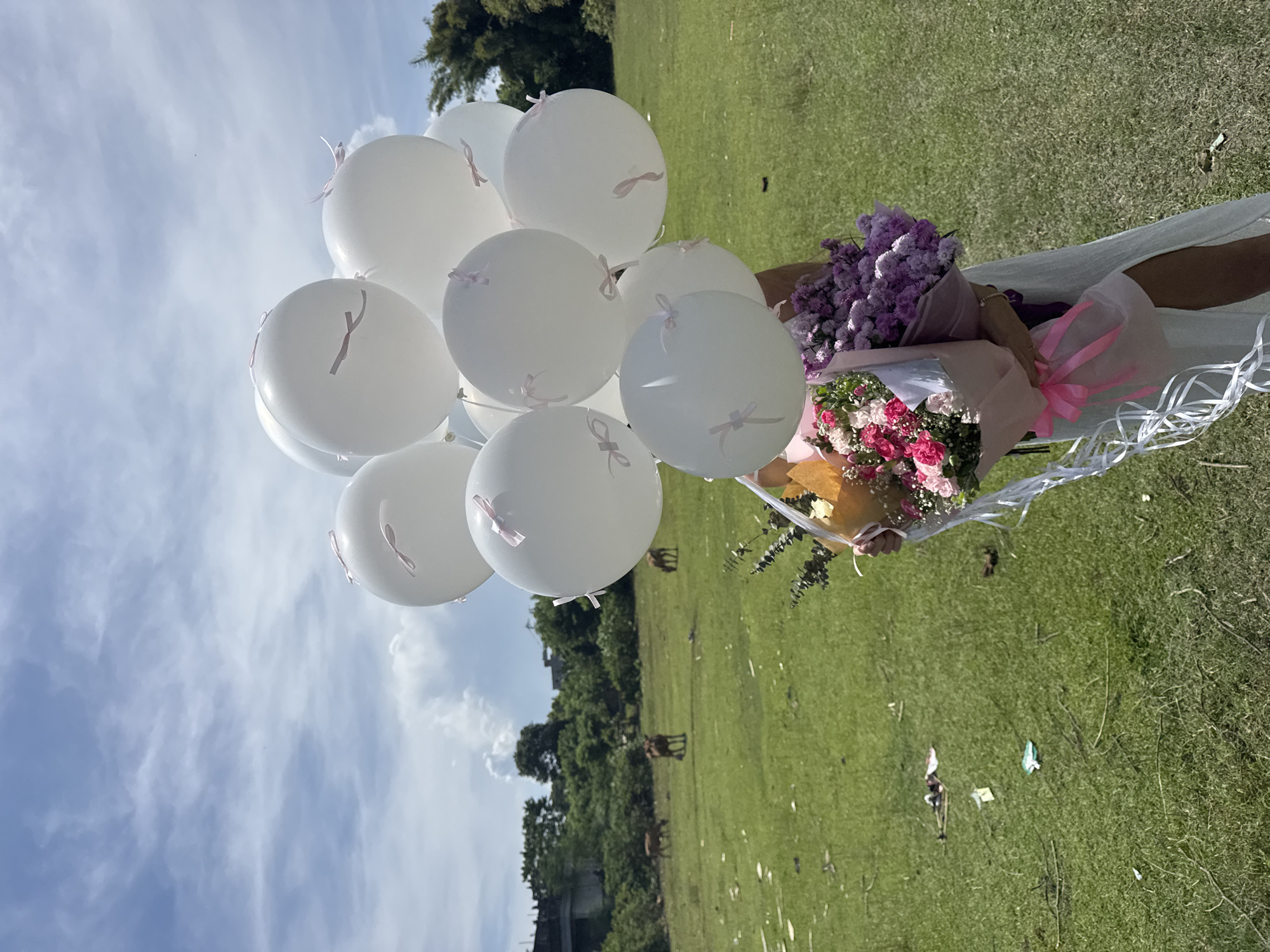 The image depicts a sunny outdoor scene in a grassy area, likely a field or park, in an unknown city. A person, holding a bouquet of flowers and a bunch of white balloons with pink ribbons, stands prominently. The bouquet consists of two wrapped flower arrangements: one with pink and white flowers, the other with purple flowers. The balloons are arranged in a cluster and partially obstruct the view of the sky, which is light blue with scattered clouds. In the background, a line of trees borders the field, with signs of human activity like scattered debris. The grass appears green and well-maintained. In the far background, there is a vague view of urban infrastructure.
