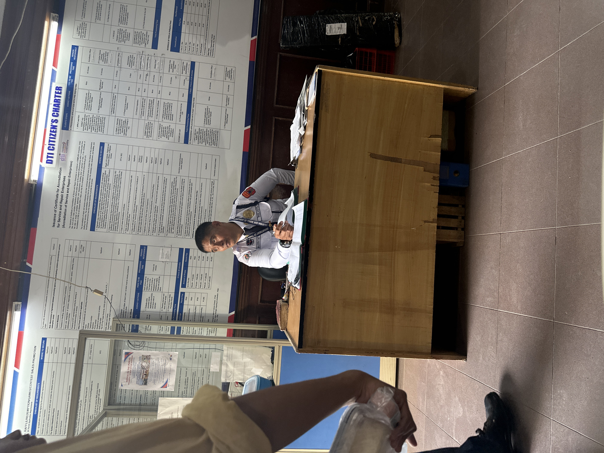 The image shows a male security guard sitting behind a wooden counter in an office. He is wearing a white uniform with patches and a badge that says "PHILIPPINES" and "MAGUINDANAO." He is looking to the side and holding a pen. There are papers and a book on the counter. Behind the security guard, a large poster with text and charts is displayed on the wall. The poster has the title "DTI CITIZEN'S CHARTER." The floor is tiled. In the foreground, a person's arm and hand are visible, holding a clear plastic bag. The scene appears to be a government office, likely the Department of Trade and Industry (DTI) in Davao City, Philippines, given the presence of DTI signage and the mention of "Davao City" in the prompt.