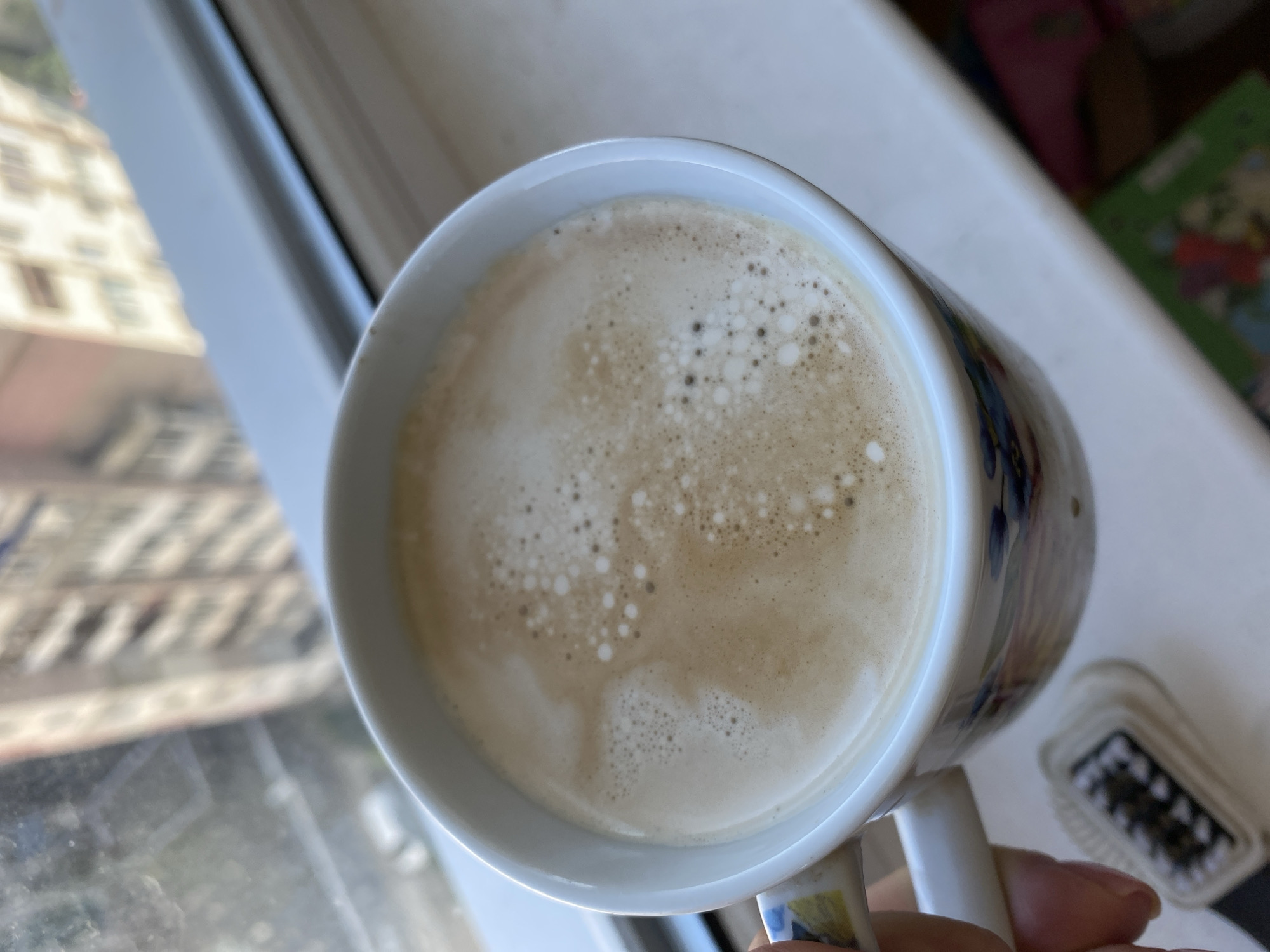 A close-up, eye-level shot shows a white ceramic mug filled with coffee and foam, held by a person's hand. The mug has a floral pattern in blue and yellow. In the foreground, to the left of the mug, is a small black and white object with bristles, possibly a brush. The background is out of focus, showing a window with a view of a city building and a window sill. The overall lighting suggests it's daytime. The image was captured in the city of Chornomorsk, Ukraine.