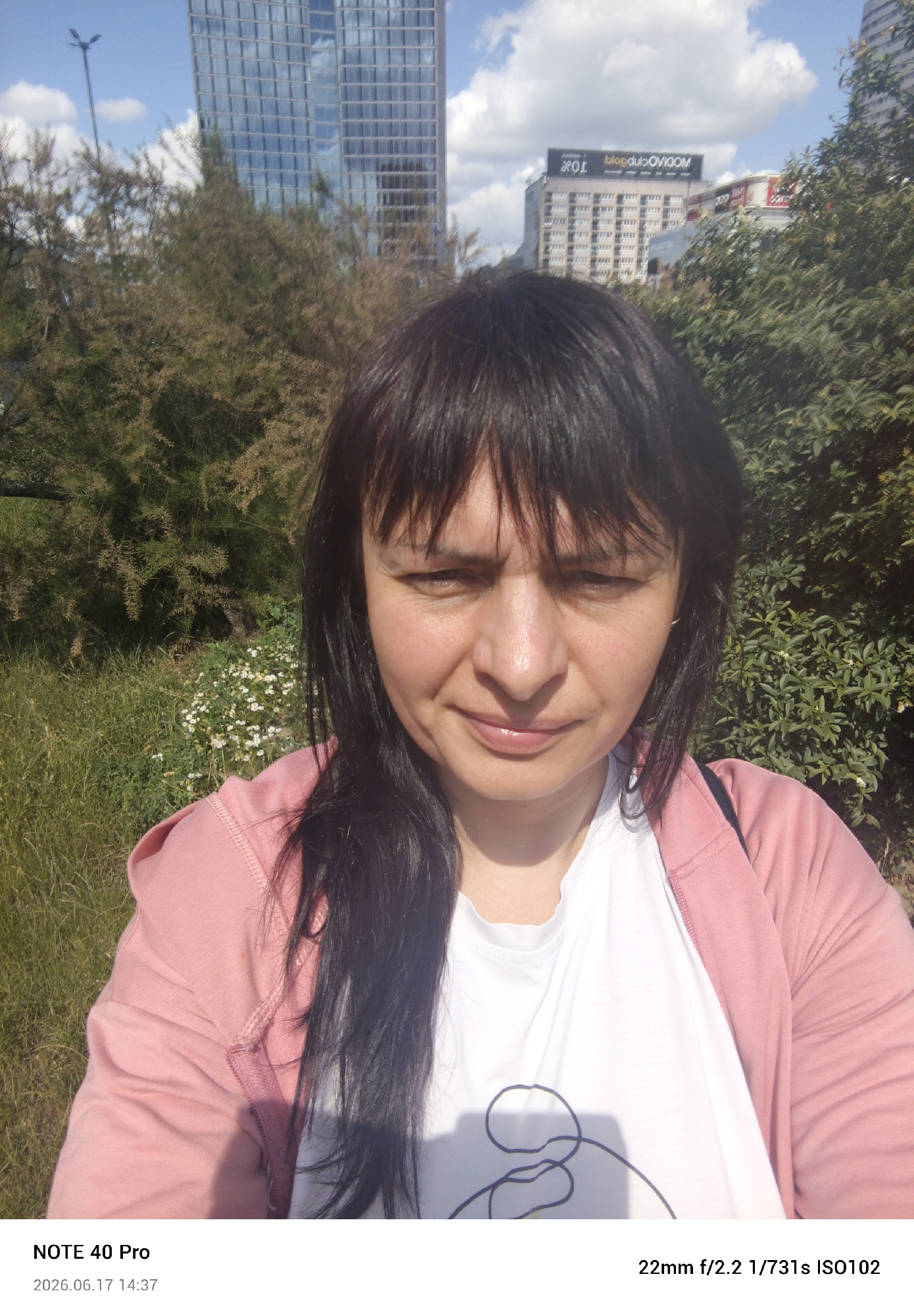 The image shows a selfie of a woman with dark hair and bangs, wearing a pink hooded jacket over a white t-shirt with a graphic design. She is outdoors with a cityscape in the background, featuring tall buildings and trees. The date and time stamp at the bottom left corner indicates "2026.06.17 14:37" and the camera model is "NOTE 40 Pro". The camera settings are also displayed as "22mm f/2.2 1/731s ISO102". The scene appears to be Warsaw, Poland, given the presence of a building with the text "blog.duolvgom.com" which is a known advertisement in Warsaw.