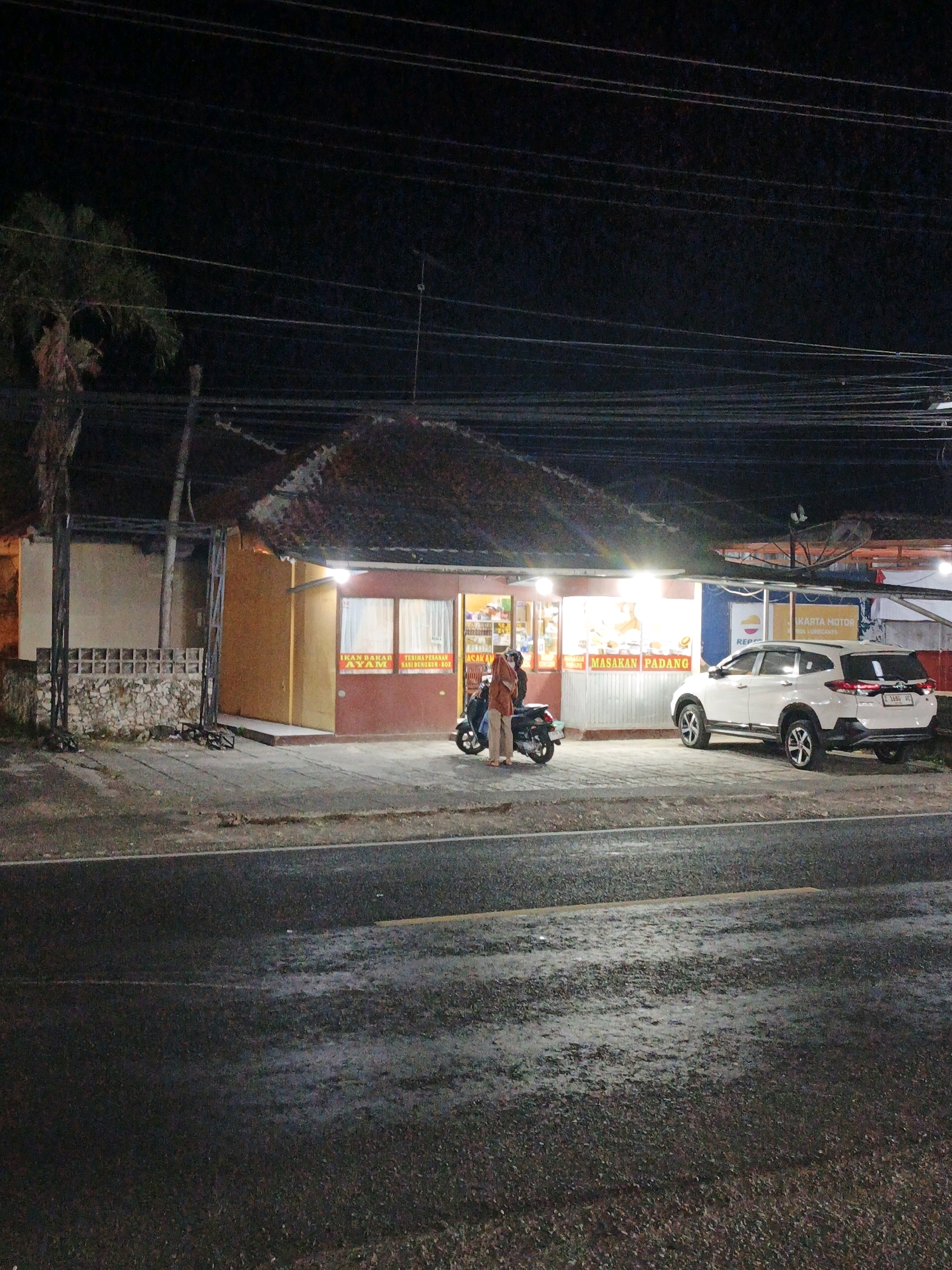 This image depicts a nighttime street scene in Pangandaran, Indonesia, specifically in front of a food establishment. A white SUV is parked to the right, facing away from the camera. To the left of the SUV, a motorcycle is parked, and a person in a reddish-brown jacket and light-colored pants is standing next to it, facing the food establishment. The food establishment has a brown awning over its roof and a warmly lit interior visible through windows. Signage on the establishment reads "IKAN BAKAR AYAM" (Grilled Fish Chicken) and "MASAKAN PADANG" (Padang Cuisine). A second person, also wearing a reddish-brown top, is partially visible inside the establishment. The street in front of the establishment appears wet, suggesting recent rain. The overall environment is dimly lit, typical of a street scene at night, with visible overhead power lines and a palm tree to the far left.