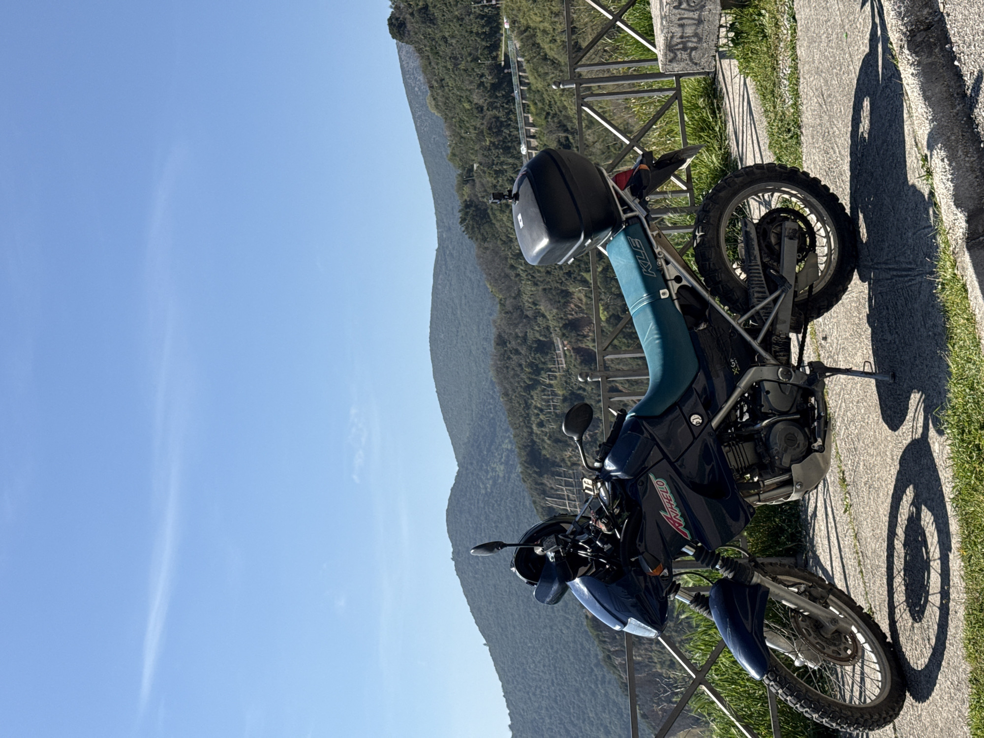 A blue Kawasaki KLR650 motorcycle is parked on the side of a paved road with a metal guardrail. In the background, a green, mountainous landscape stretches under a clear, blue sky. The motorcycle is facing left, with its shadow cast on the pavement. The location is Prijevor, Montenegro, suggested by the mountainous landscape and probable road infrastructure.
