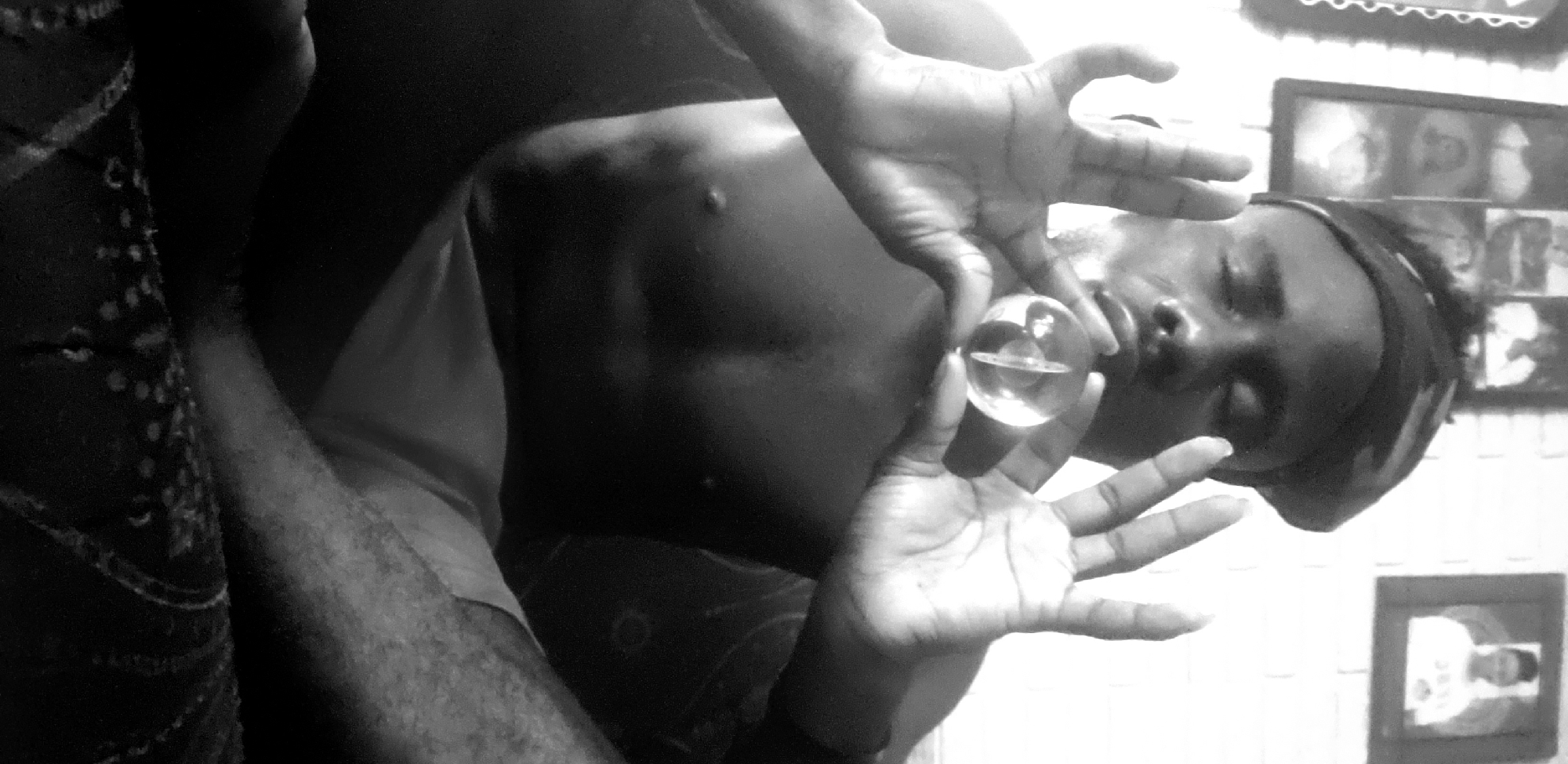 A shirtless person with a headband holds a glass ball in front of their mouth with both hands. The person is in an indoor environment with framed photos visible on a wall. Based on the prompt, the location is Narayi, Nigeria.