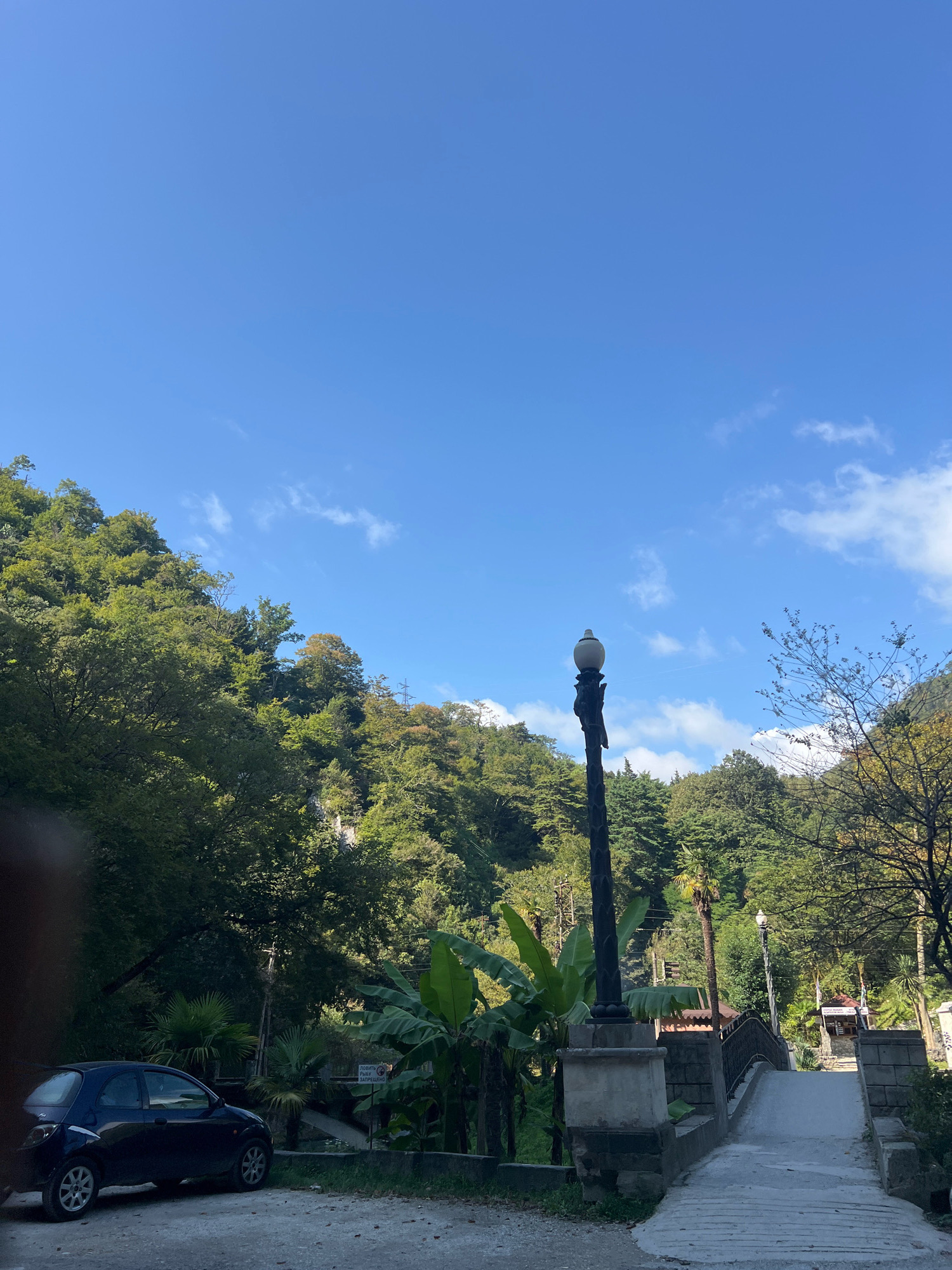 A dark blue hatchback car is parked on a dirt and gravel surface. The car is facing left, with its front end obscured by the edge of the frame. Behind the car, lush green trees and foliage cover a steep hillside. A narrow, concrete path leads uphill towards a small stone bridge. The bridge has an ornate black metal railing. To the left of the path and bridge stands a tall, dark lamppost with a white globe light. Further up the hill, a small building with a red roof is visible, and a tall evergreen tree stands to the right of the bridge. The sky is a clear, bright blue with scattered white clouds. The scene appears to be a park or natural area in a mountainous region. The location is identified as New Athos, Abkhazia, suggesting a resort or tourist destination.