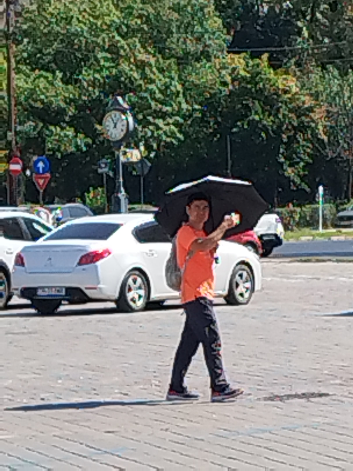 A man is walking on a paved street in Bucharest, Romania, holding a black umbrella over his head and a colorful object in his right hand. He is wearing an orange t-shirt and dark pants. Several white cars are parked in the background. A large clock is visible on a pole in the upper left corner of the image. The sky is clear and the scene appears to be illuminated by sunlight.