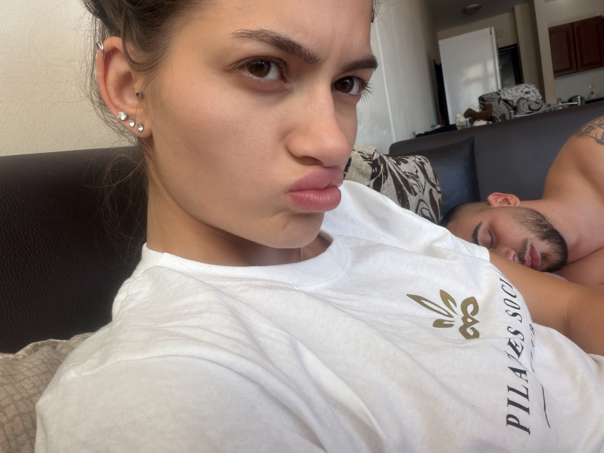 A woman with dark hair pulled back is posing for a selfie, pouting her lips and looking directly at the camera. She is wearing a white t-shirt with a gold logo and text that appears to read "PILATES SOCIAL." She has multiple piercings in her left ear. To her right, a man is lying down, asleep, partially visible. He has dark hair and a beard. They are both on a dark colored couch with patterned pillows. The background shows a glimpse of an interior space with cabinets and a refrigerator, suggesting a home environment. The lighting is natural and appears to be daytime.