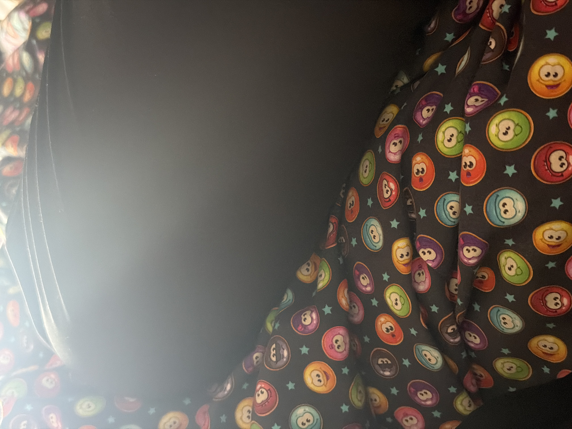 The image presents a close-up, detail-oriented view of patterned fabric against a plain, dark background. The dominant feature is a dark-colored fabric, likely black or very dark gray, adorned with a repeating pattern of colorful, stylized oval or circular shapes. These shapes, resembling cartoon characters or pool balls, display simple, cheerful faces with wide eyes and varied expressions. They come in a palette of bright colors including red, orange, yellow, green, light blue, purple, and pink. Many of these shapes feature additional markings inside, such as the number "8" (visible on some green and blue shapes) or the letter "B" (visible on some red shapes), reminiscent of billiards. Small, light blue, five-pointed stars are scattered throughout the pattern.
The fabric appears to be rumpled or draped, suggesting it might be an article of clothing or upholstery. A significant portion of the image, particularly the upper left and central area, is occupied by a plain, dark, unpatterned surface, which could be another piece of solid-colored fabric or a dark object. A bright light source or glare is visible in the lower-left quadrant, creating a soft, hazy glow over a segment of the dark area and the patterned fabric. No people are visible, nor is there any discernible activity taking place. The overall impression is one of a playful or whimsical design, captured in an indeterminate indoor setting. No specific location cues for Iskar, Bulgaria, are visible.