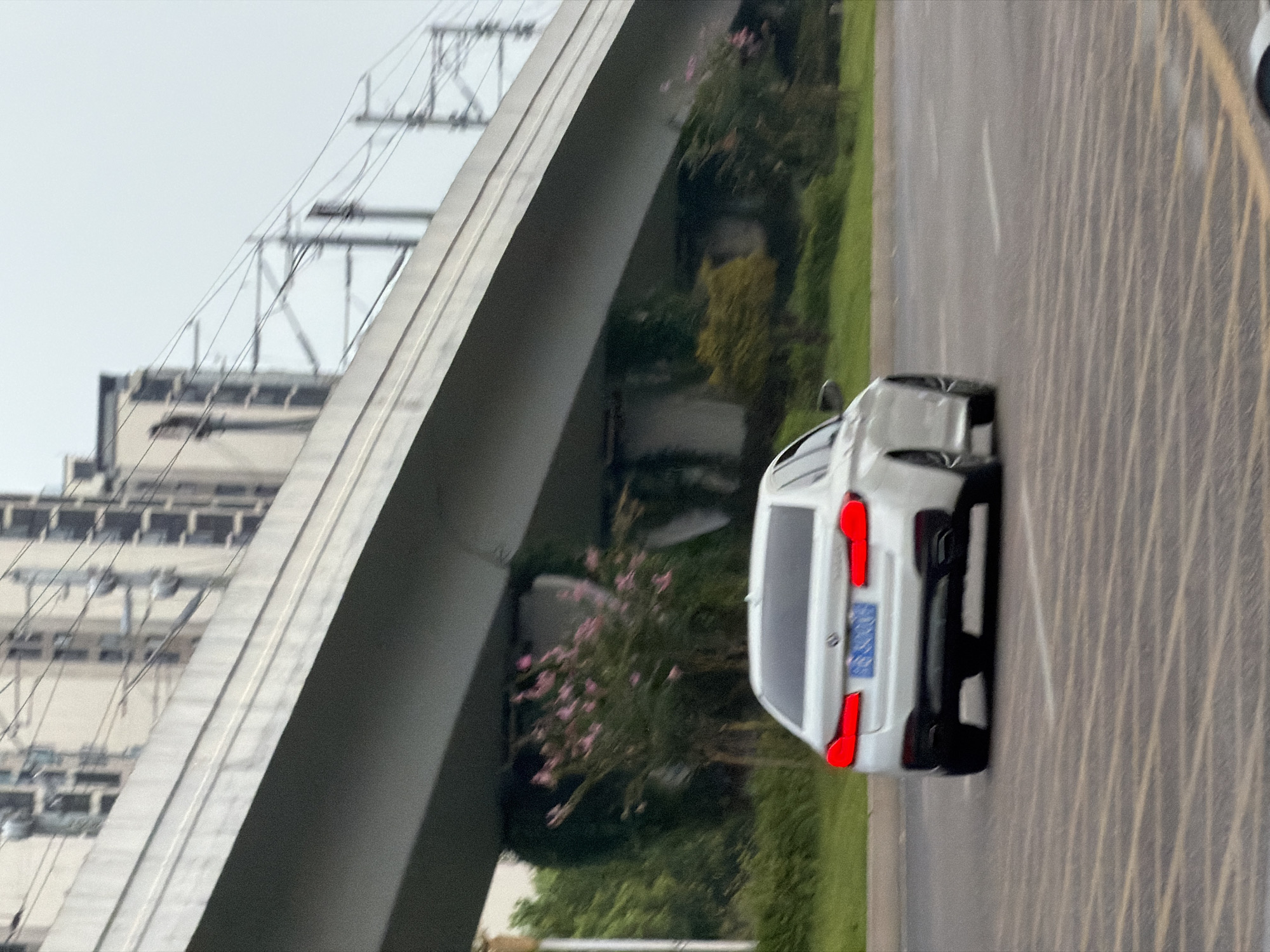 A white BMW sedan is traveling on a paved road under an elevated structure, likely a highway or train track. The car's rear taillights are illuminated. The road surface features distinct yellow and white striped markings. In the background, there are buildings and vegetation, including flowering trees with pink blossoms. The sky is overcast. The license plate of the car, though blurred, appears to be blue with white characters, consistent with Chinese license plates. The scene is captured from a slightly low angle, looking towards the rear of the moving vehicle.