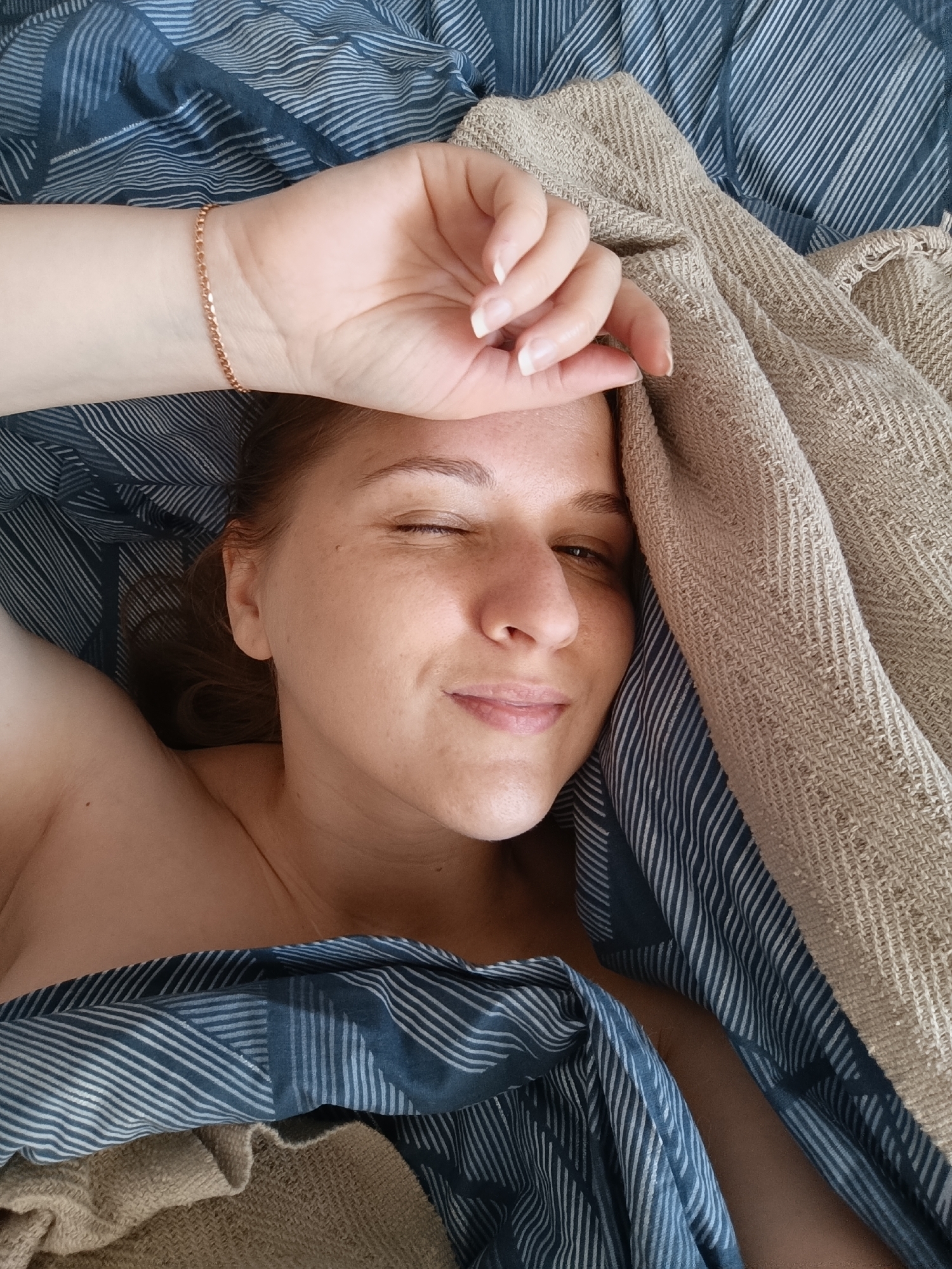 A woman with light skin and brown hair is lying in bed, partially covered by blankets. Her right hand is raised to her forehead, and she is winking at the camera. The blanket covering her has a pattern of blue and white stripes, and another blanket, a beige textured fabric, is draped over her. A thin, rose gold bracelet is visible on her wrist. She appears to be in a relaxed or playful mood. The setting appears to be a bedroom.