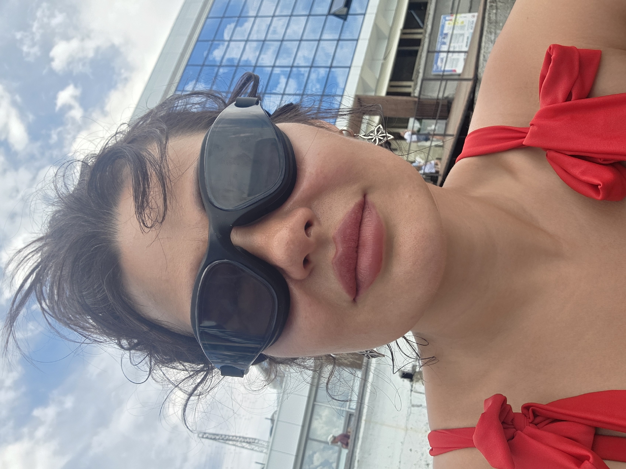 A woman wearing swimming goggles and a red swimsuit is pictured close-up, with a blue glass building and cloudy sky in the background. She has dark, messy hair and is wearing ornate silver earrings. The image appears to have been taken outdoors, possibly near a body of water, given the swimming goggles and swimsuit. The architectural style of the building suggests a modern urban setting, and the overall atmosphere is relaxed and casual. The specific location within the city of Sochi, Russia, cannot be determined from the visual information alone, but the presence of a modern building and what appears to be a waterfront promenade points towards a developed area.