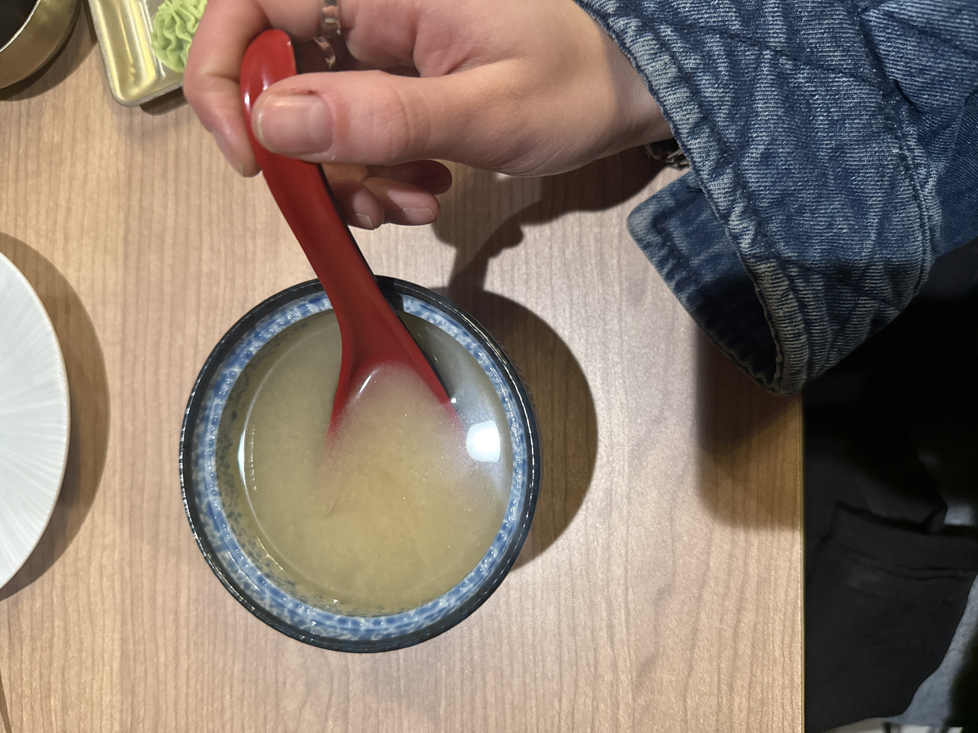 Here is a detailed description of the image:
**Scene:** The image is a close-up, overhead shot of a dining setting.
**Objects:** A bowl of miso soup with a red plastic spoon inside is the central focus. Beside the soup bowl is a small white plate and a container of what appears to be wasabi.
**People:** A person's hand is holding the red spoon, about to stir or taste the soup. The individual is wearing a denim jacket.
**Environment:** The setting is a wooden table, likely inside a restaurant in Southlake, United States of America. Black pants can be seen off the right side of the photo.