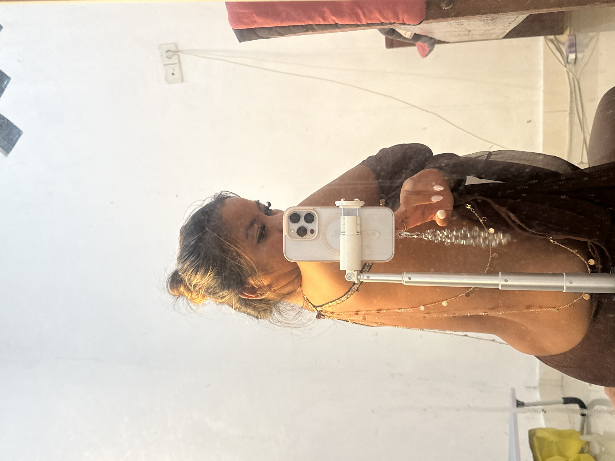 A selfie of a woman in a mirror reflection in Celuk, Indonesia. She holds a white iPhone with a tripod stand, obscuring part of her face. She wears a brown top and multiple layered necklaces. Her hair is tied up. The background includes a white wall with an electrical outlet, a shelf with pink clothing, and the corner of a white shelf.