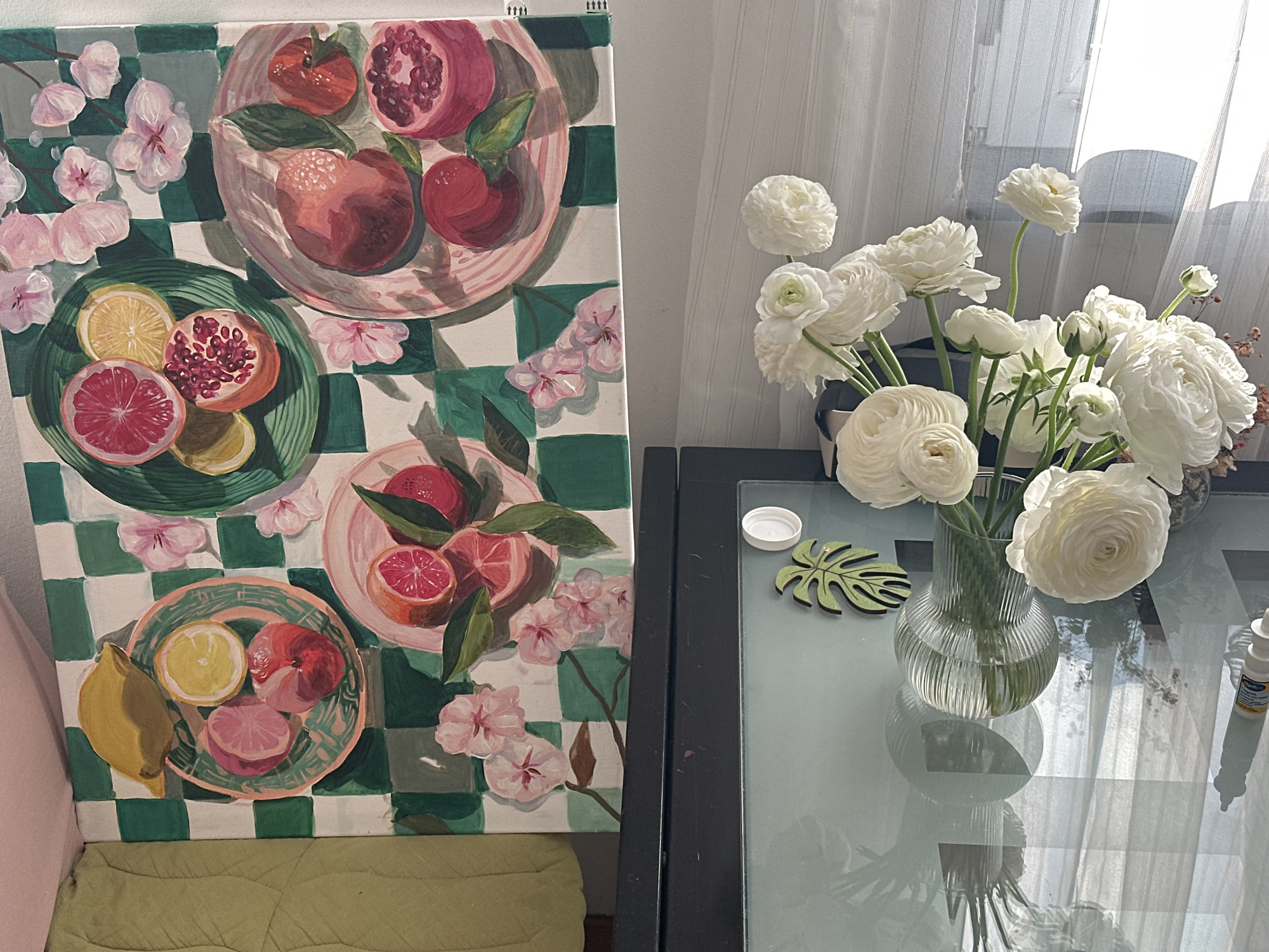 A still life is depicted, featuring a painting of a fruit and flower arrangement positioned next to a vase of white ranunculus flowers. The painting displays several plates of fruit, including citrus and pomegranates, set against a green and white checkered background, with pink flowers interspersed. The vase containing the flowers is clear and sits on a glass table. A small, green, leaf-shaped coaster and a white bottle are also on the table. The environment appears to be indoors, with a sheer curtain in the background, indicating a domestic setting. It is not possible to confirm the specific location in Novi Sad, Serbia, based on the image provided.