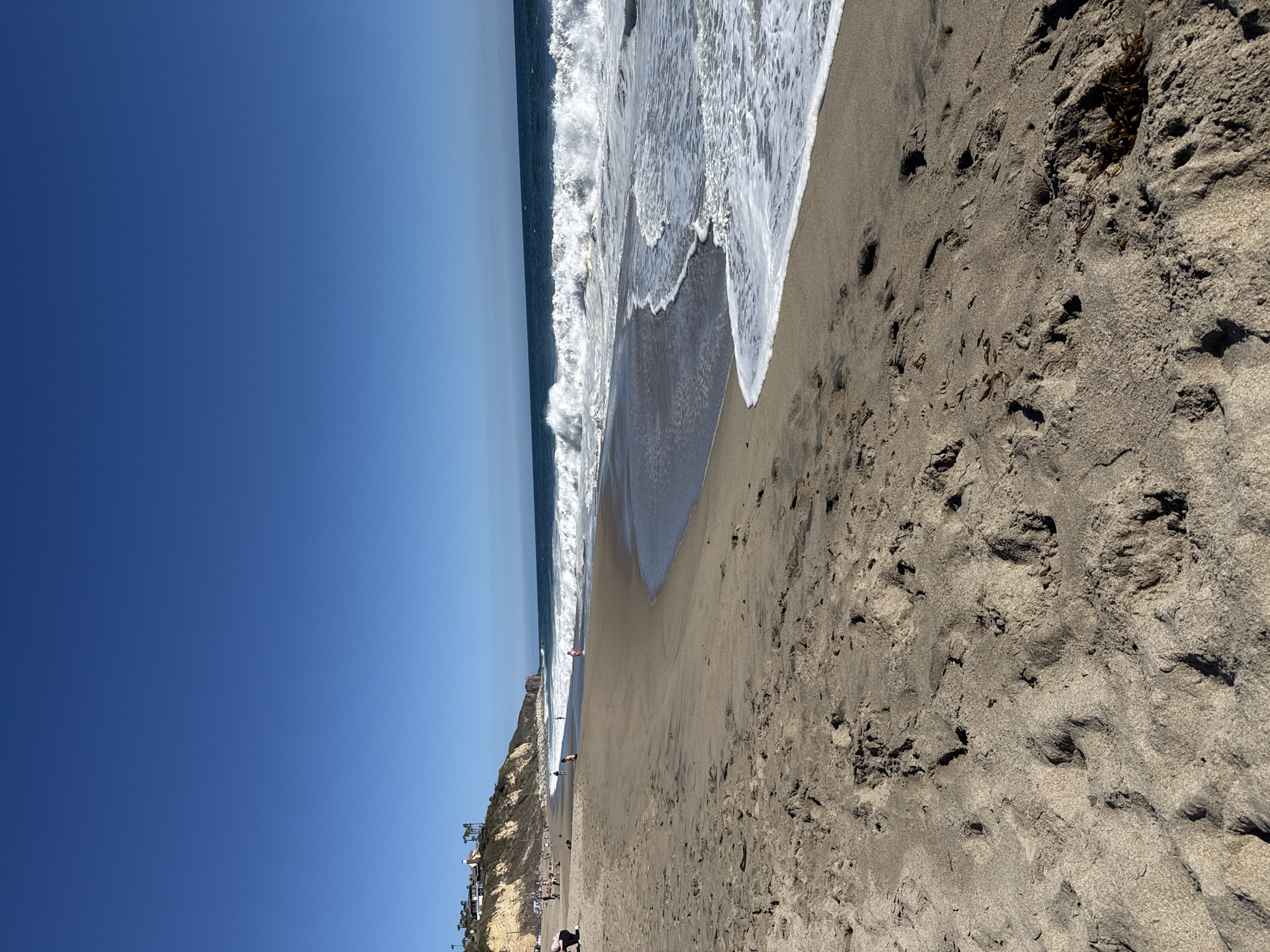 The image depicts a wide, sandy beach in Malibu, California, United States of America, on a clear, sunny day. The foreground is dominated by the textured sand, showing numerous footprints and some scattered seaweed. The ocean, with its characteristic blue hue, occupies the middle ground, featuring moderately sized waves breaking on the shore, creating white foam. In the background, a rugged cliff face rises, topped with houses and palm trees, indicative of a coastal residential area. Several people are scattered across the beach, some walking, others sitting or playing near the water's edge. The sky is a deep, cloudless blue.