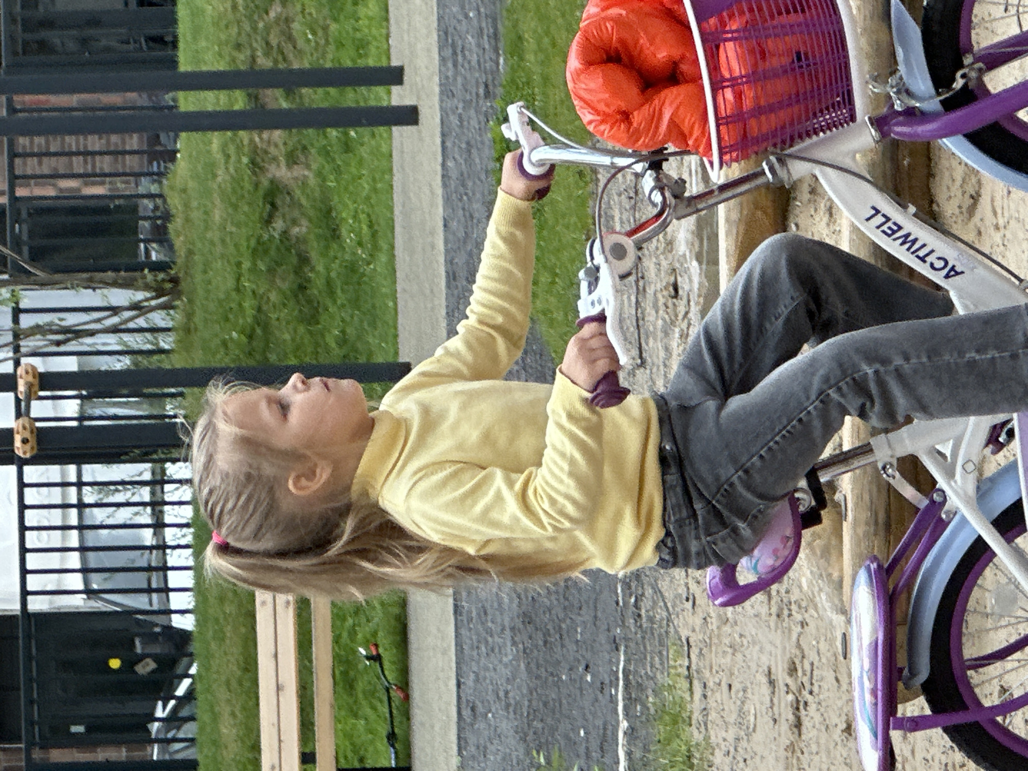 A young girl with blonde hair in a ponytail, wearing a yellow turtleneck sweater and dark jeans, rides a white bicycle with purple accents. The bicycle has a purple basket in front filled with orange items, possibly toys or clothing. The girl is on a sandy playground with a wooden bench and a fence in the background. The environment appears to be a park or a residential area in Znamya Oktyabrya, Russia, with green grass and trees visible.