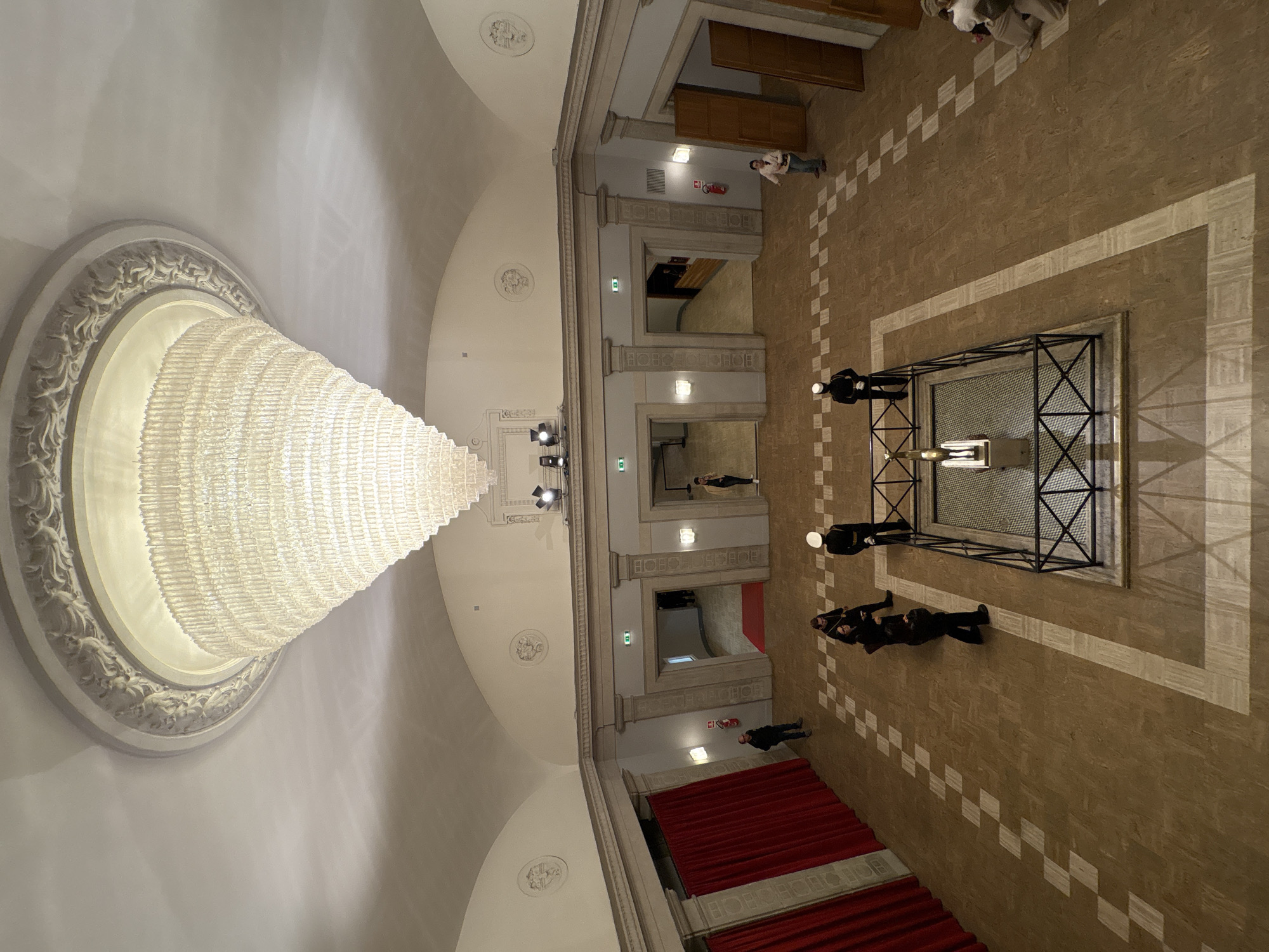 The image shows an interior space, likely a grand hall or public building in Rome, Italy. The camera's perspective is from a high vantage point, possibly an upper level, looking down.
A large, ornate chandelier dominates the left side of the frame, casting light onto the surrounding ceiling adorned with classical plasterwork.
The floor is tiled in a neutral tone with a decorative checkerboard pattern. Several doorways line one side of the hall, suggesting a series of interconnected rooms or spaces. Red carpeting is visible near the bottom of the frame, possibly indicating a formal area like a stage or seating section.
Several individuals are present in the scene. Some appear to be standing or walking near the doorways, while a group is gathered around what appears to be a monument or cenotaph enclosed by a dark metal framework. The people near the monument seem to be in a formal posture, possibly paying respect or participating in a ceremony. The general setting conveys a sense of formality and solemnity.