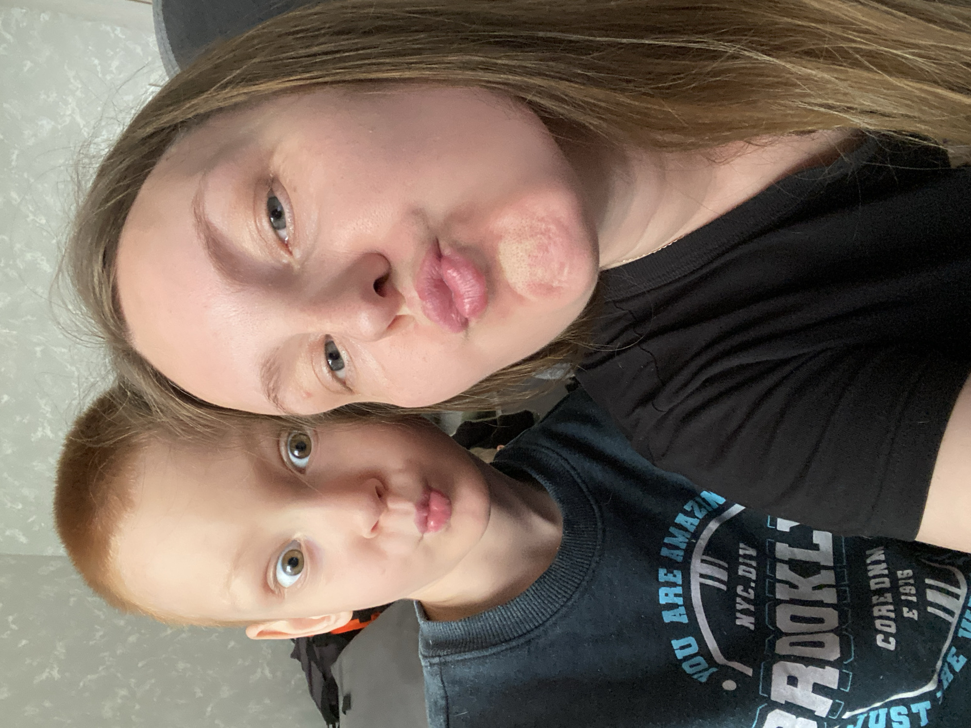 A selfie features a woman and a young boy, positioned closely together and looking at the camera with pursed lips. The woman, who is older and light-skinned, has brown hair and is wearing a black t-shirt. The boy has red hair and wears a dark blue sweatshirt with the word "BROOKLYN" on it. The background is a neutral gray wall. There is no evidence to suggest the location is Noviy, Russia.