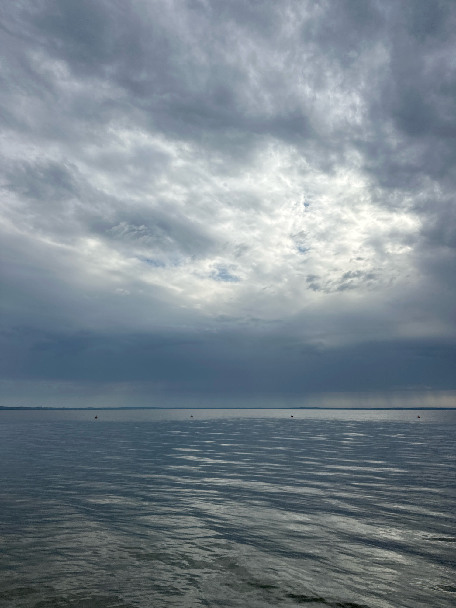 The image shows a wide expanse of water under a cloudy sky. The water is calm with gentle ripples, reflecting the muted light from the overcast sky. On the horizon, a faint line of land is visible. There are two small, red buoys in the water, positioned at a distance from each other. The sky is filled with dramatic, heavy clouds, with patches of brighter light breaking through in the upper portion of the frame. The overall mood is somber and tranquil. This scene is likely located on Lake Naroch, within the Naratski Selsoviet, Belarus, known for its expansive water bodies and natural landscapes.