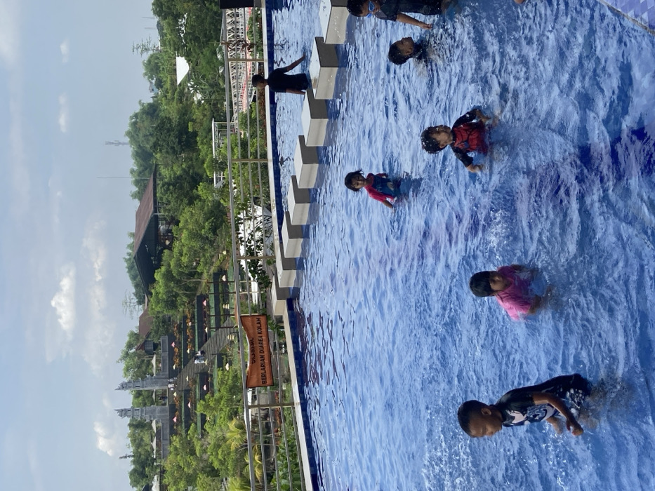 Children play in a pool on a sunny day. The pool has a row of rectangular steps along one side. Beyond the pool and a metal railing, lush green trees fill the landscape. Traditional Indonesian architecture is visible among the trees. The sky is partly cloudy.