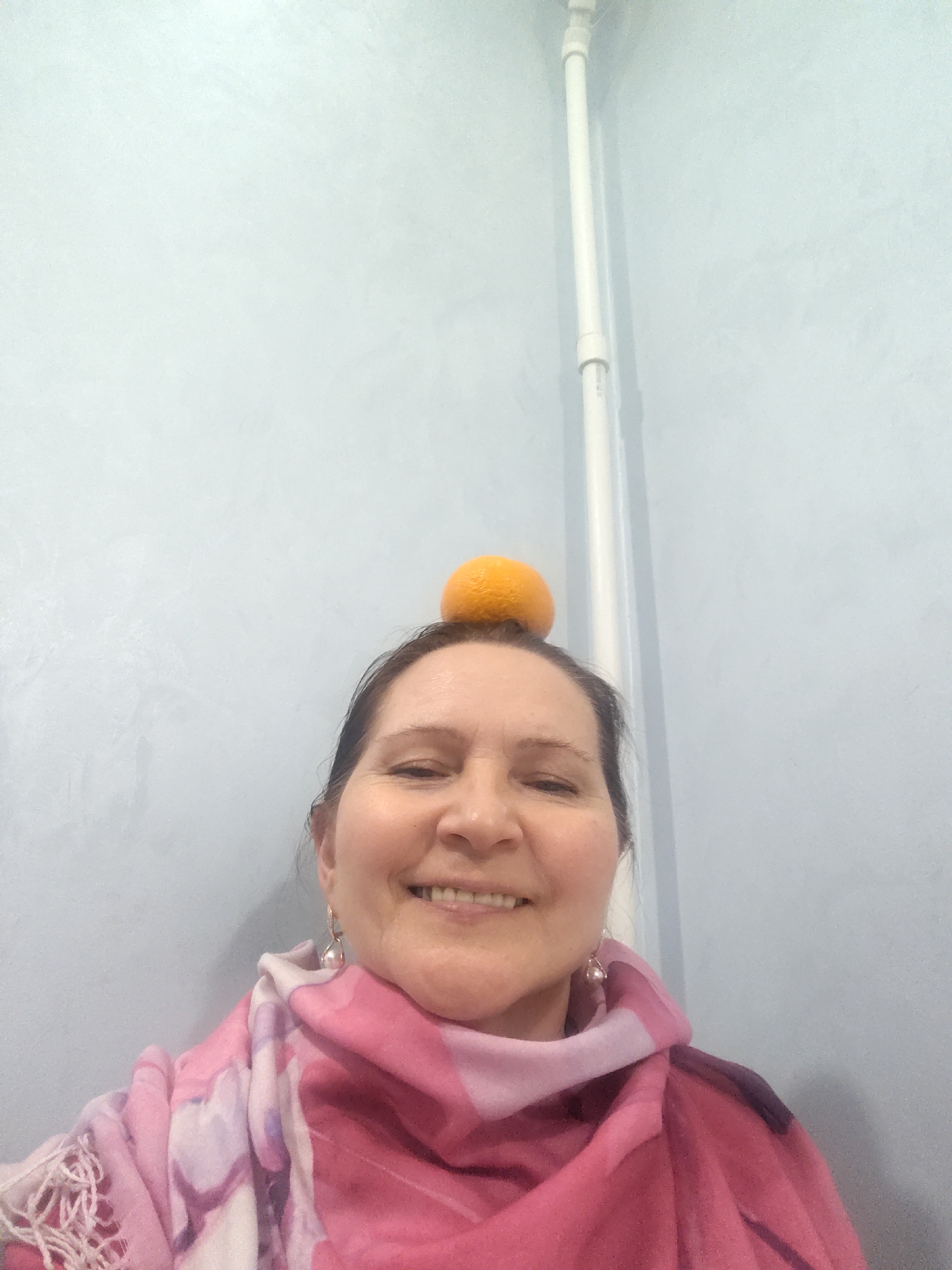 The image shows a middle-aged woman with fair skin, brown hair, and a slight smile. She is wearing a pink and white patterned scarf. An orange is balanced on her head. In the background is a light blue wall with a white pipe running vertically. The location in the city of Sarapul, Russia is not discernible from the provided image.