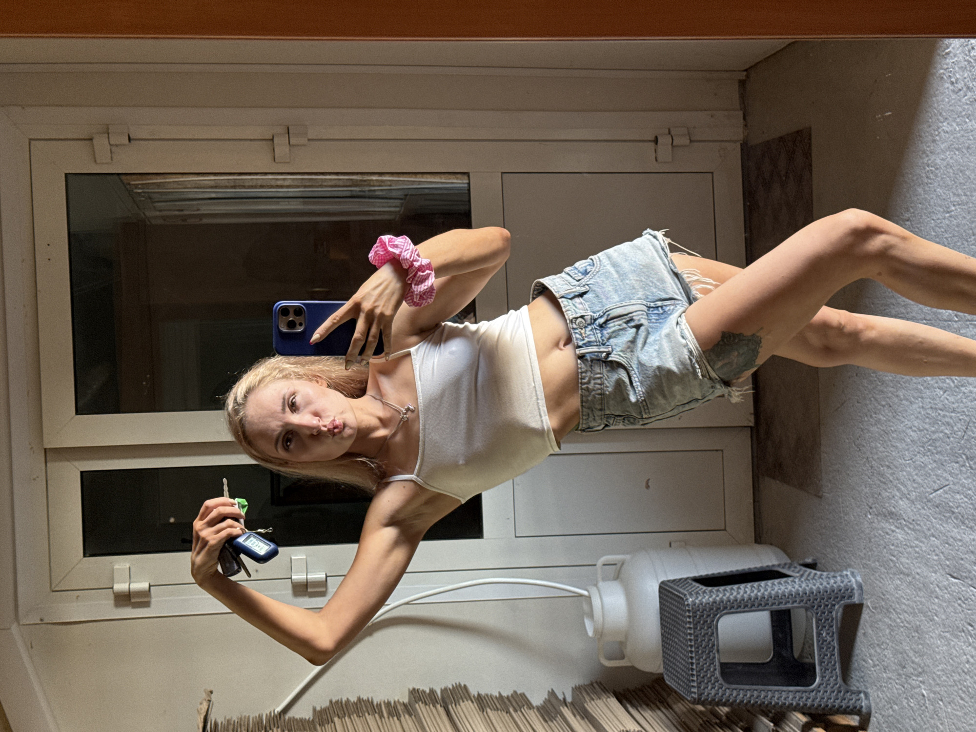 The image shows a young woman taking a selfie in what appears to be a hallway or entryway. She is holding a blue smartphone in her right hand and a set of keys in her left. She is wearing a white tank top and denim shorts. Her hair is blonde and pulled back with a pink and white striped scrunchie. She has a necklace on. The background features a door with a window, a water cooler, and a small gray plastic stool. The floor is gray and appears to be made of concrete.
Based on the available information, it is not possible to confirm the location in Chaikovsky, Russia. The information does not contain location specific landmarks. The scene shows details typically found in a hallway.
