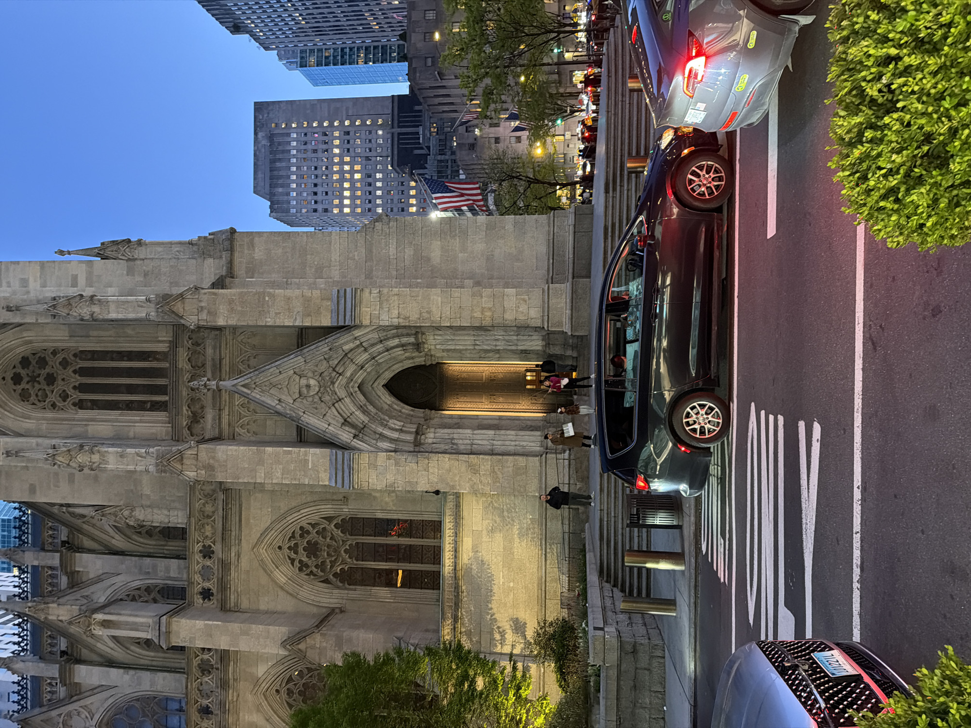 The image shows a street view in New York City, featuring St. Patrick's Cathedral on the left. The cathedral's stone façade is visible, with its gothic architectural details, including arched windows and intricate carvings. People are standing on the steps of the cathedral's entrance.
On the right side of the image, a city street is visible. Cars are parked along the curb. Street markings are visible on the asphalt.
In the background, skyscrapers are seen, indicating the urban setting. The sky is a deep blue, suggesting it is either dusk or dawn. An American flag is prominently displayed, and several other flags are visible further down the street. The overall lighting indicates the scene is captured during the early evening or late afternoon.