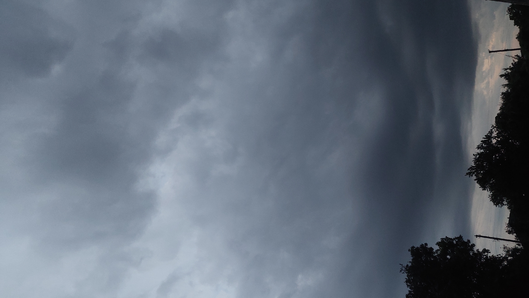 The image depicts a dark, cloudy sky over Kozarovychi, Ukraine. Silhouetted trees and utility poles are visible against the ominous sky in the lower portion of the frame. The clouds are dense and appear to be heavy, suggesting impending rain or a storm. The overall atmosphere is somber and dramatic.