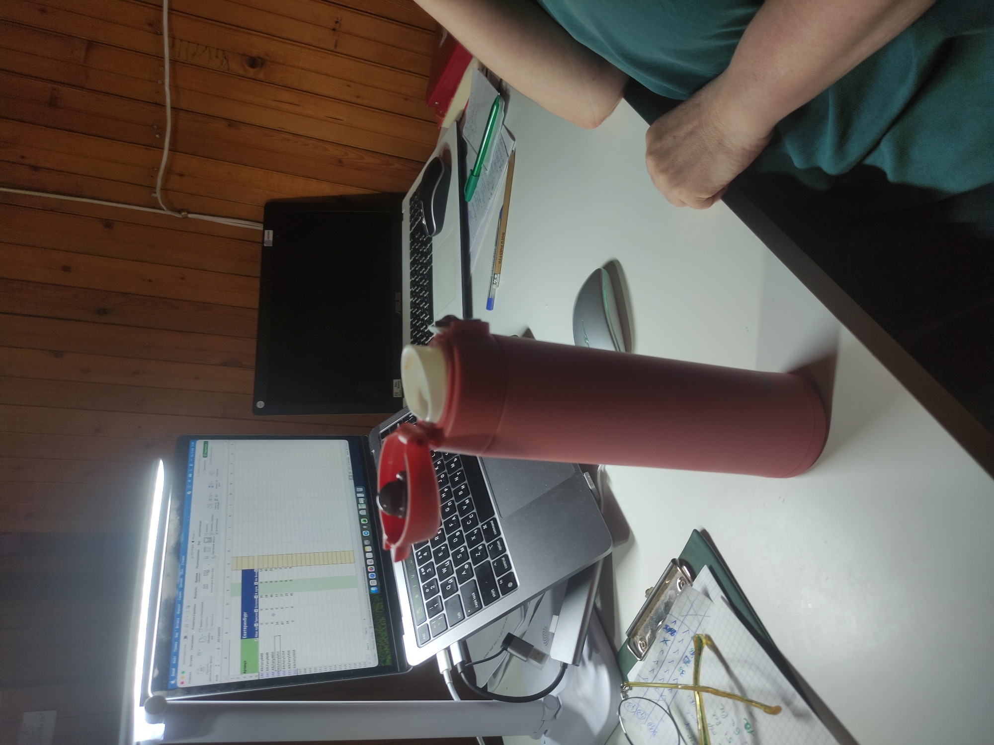 A desk scene shows two open laptops displaying spreadsheet data, a pink thermos, and eyeglasses lying on a clipboard. The desk surface is white, positioned against a wood paneled wall. The location and details of the city are unknown. A person's arm is visible above the desk, wearing a green shirt. The overall scene suggests a workspace, possibly a home office.