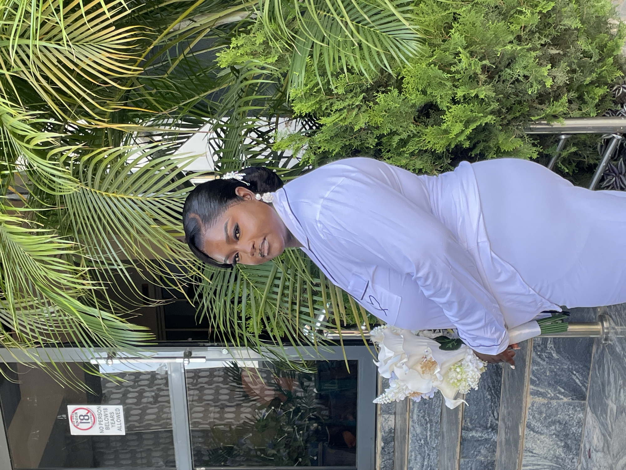 A medium shot depicts a woman in Jos, Nigeria, holding a white floral bouquet while standing on grey steps outside a building. She wears a long-sleeved white garment with a pocket detail. Behind her are dense green plants, including palm fronds. A glass door is visible, displaying a sign that reads "NO PERSON BELOW 18 YEARS ALLOWED" in red lettering. The woman gazes toward the camera. The surrounding environment consists of greenery and architectural elements.