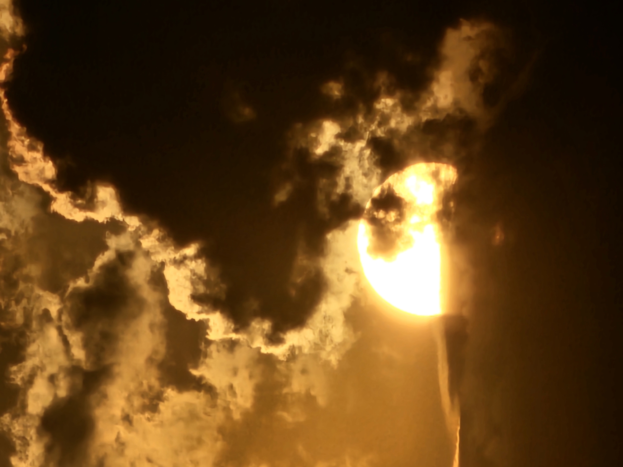 The image shows a dramatic sunset over the city of Denpasar, Indonesia. The sun, a bright, glowing orb, is partially obscured by dark, heavy clouds. The clouds are illuminated from behind by the sun, creating a halo of golden light around them. The overall impression is one of intense, fiery beauty, with the sun's rays struggling to break through the thick cloud cover. The scene is serene and awe-inspiring, capturing the power and majesty of nature.
