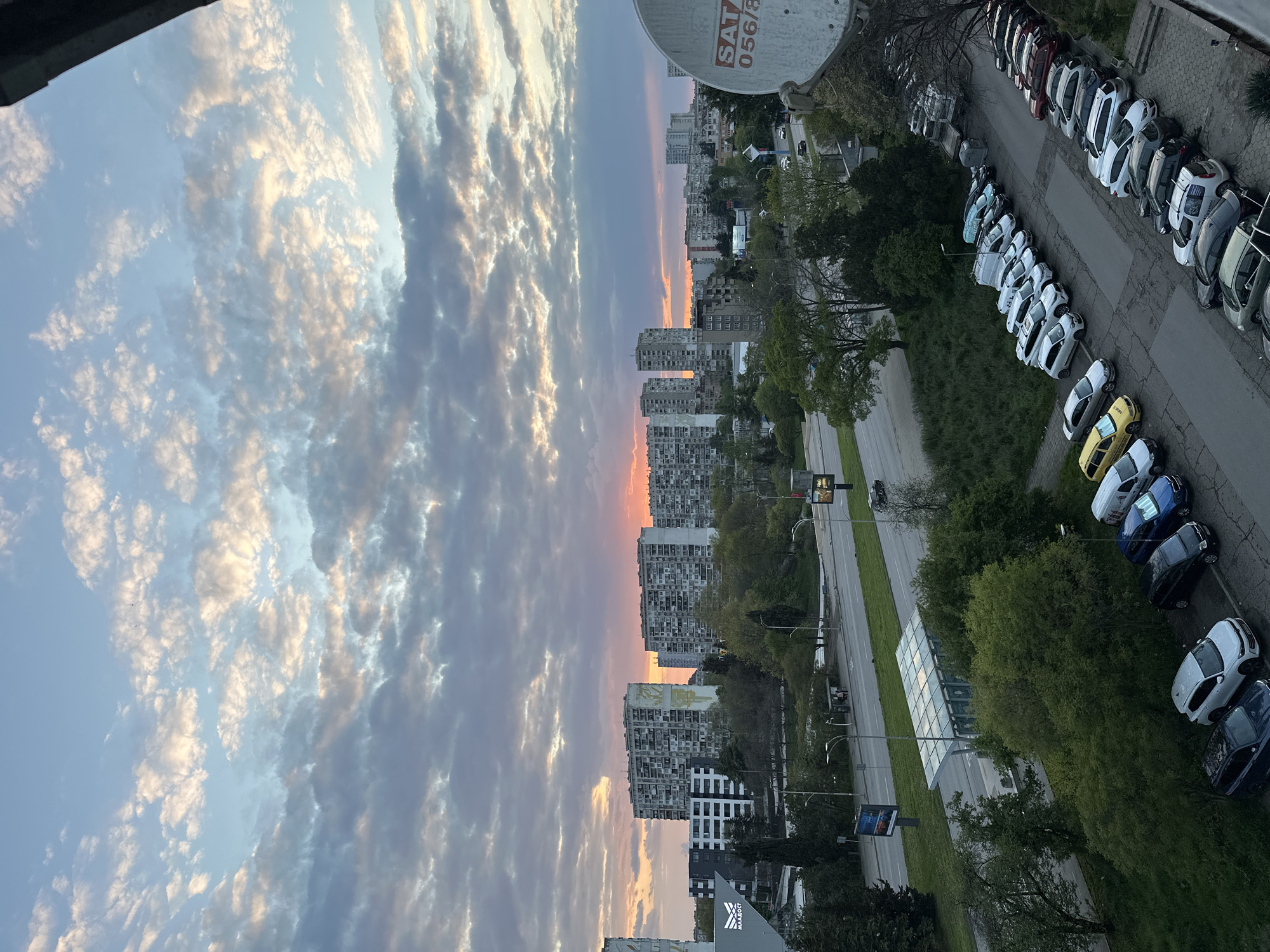 The image presents an expansive urban landscape during either sunrise or sunset, dominated by a dynamic sky. The sky is filled with numerous clouds, tinted with soft orange, pink, and yellow light near the horizon, gradually fading into lighter blues and grays higher up.
Below the horizon, a dense city sprawls, characterized by multiple high-rise residential buildings. These buildings appear to be multi-story concrete structures, some showing signs of age, while others feature more modern facades. A distinctive, large, light-colored mural is visible on the side of one building. Another building prominently displays the Cyrillic text "МААДОСТ" on its facade.
In the foreground and mid-ground, a multi-lane road traverses the scene, bordered by green grass and trees. Numerous cars are parked in organized rows along the road and in parking areas. Many vehicles are white, alongside several yellow and blue cars, suggesting ride-sharing services. A few cars are in motion on the main road, and a lone cyclist can be faintly seen. Urban infrastructure includes traffic lights, streetlights, advertising billboards, and a modern bus stop featuring a glass roof. A large white satellite dish, marked with "SAT 056/8," occupies the upper right portion of the frame. The scene conveys the quiet activity of a city during the transitional hours of the day, under a beautifully illuminated sky.