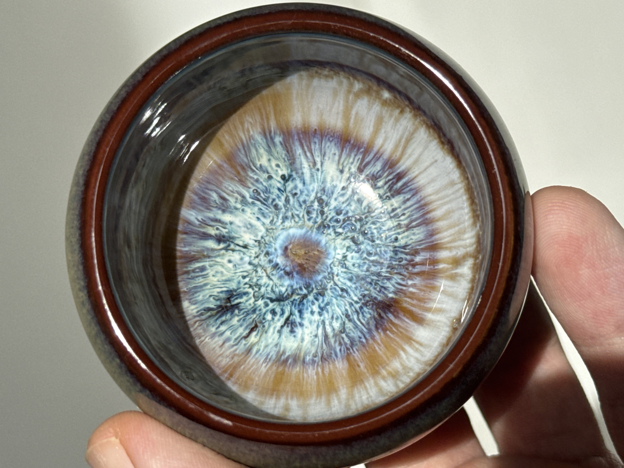 A close-up, overhead shot shows a hand holding a ceramic bowl. The bowl has a rich, glossy brown rim and the interior features a mesmerizing glaze pattern of swirling blues, whites, and golden-brown hues, resembling an eye or a nebula. The ceramic surface is slightly wet, reflecting the light. The background is a plain, light-colored surface, likely a table or countertop. No specific location in Tay Nha Trang Ward, Vietnam, is discernible from the image.