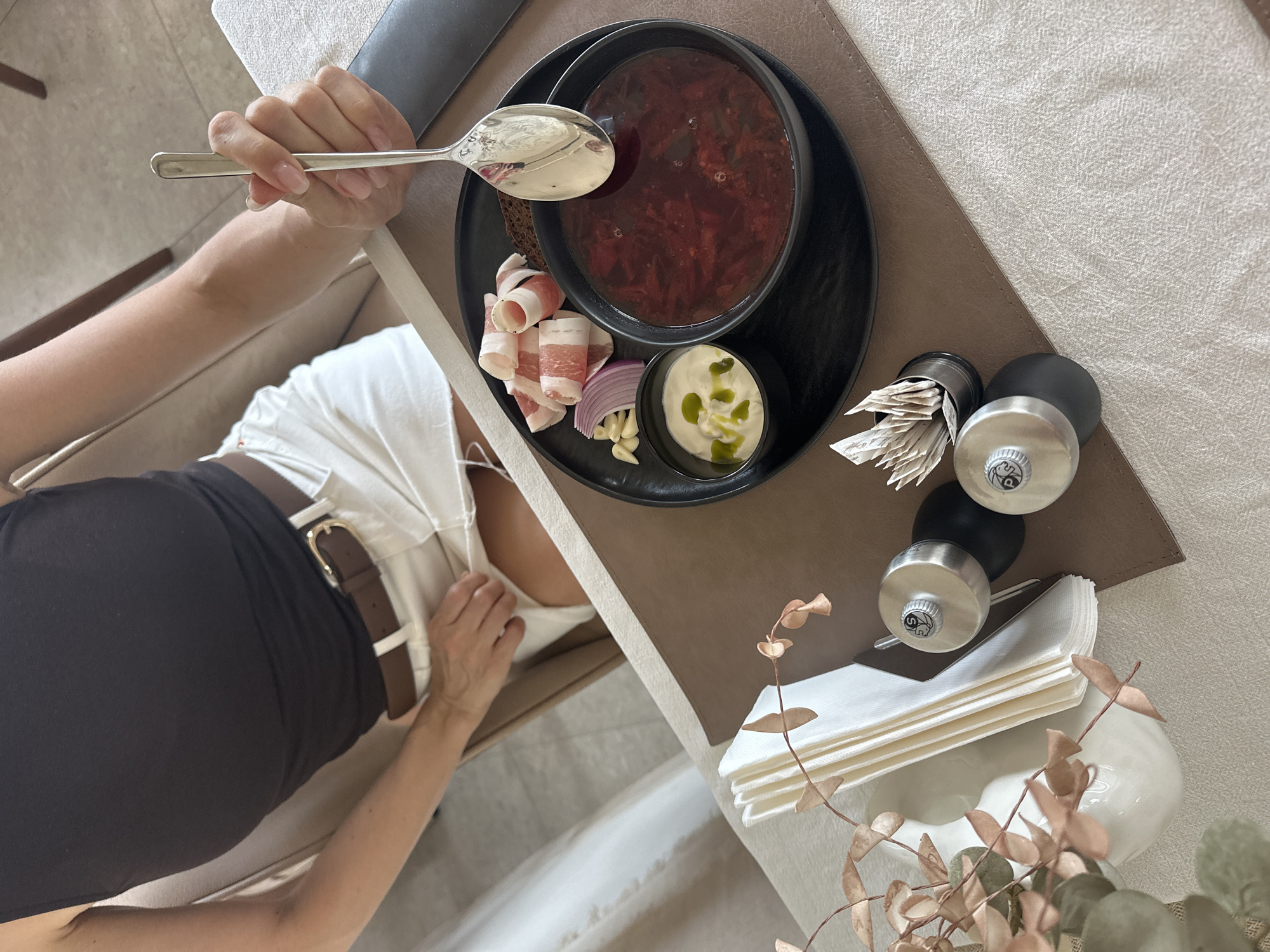 A top-down view of a person holding a spoon over a bowl of borscht on a table. The table is covered with a linen cloth, a brown placemat, and a black plate holding the borscht, bread, bacon, red onion, garlic cloves, and a small bowl of white sauce with green herbs. Next to the plate are toothpicks, salt and pepper shakers, and a stack of napkins. The person wearing a black skirt and a white top is holding a silver spoon. The location cannot be determined from the information given.