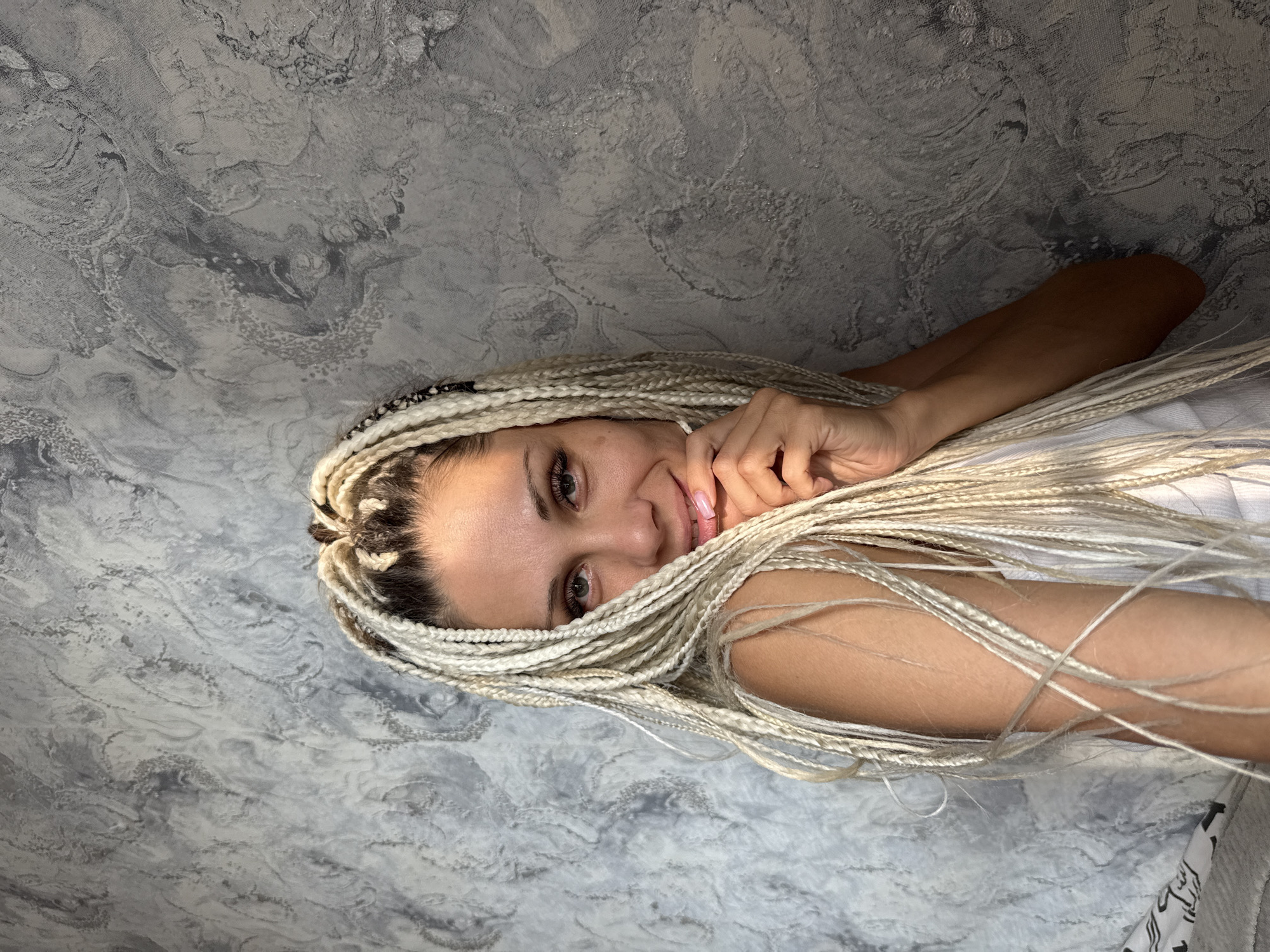 A Caucasian woman with blonde braided hair smiles and touches her face with her hand, looking at the camera. She is wearing a white top. The background is a textured grey wall. The image is a medium shot, with the woman occupying the right side of the frame. No specific location in Rusanovo, Russia is visible.