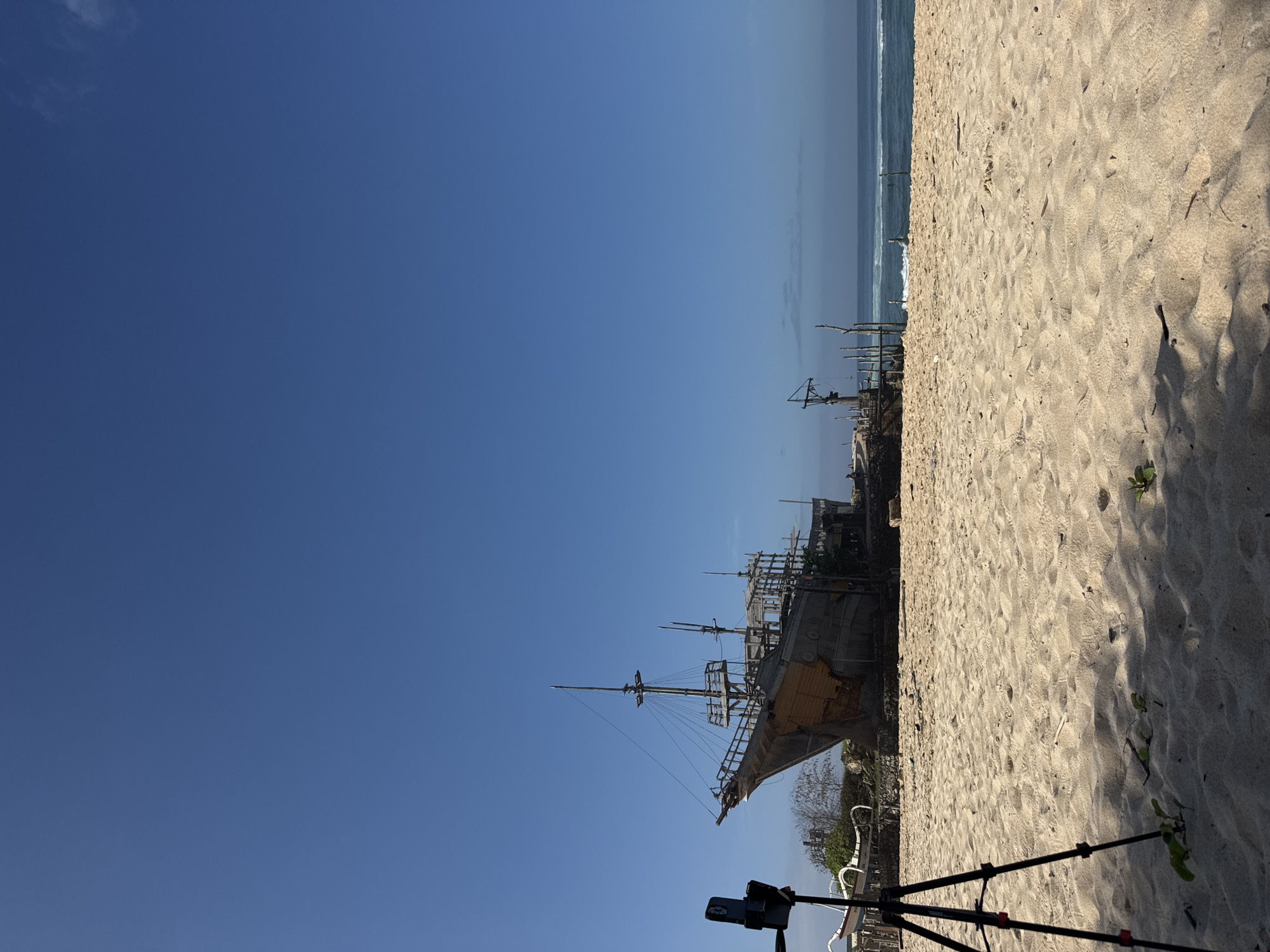 The image shows a scene at Melasti Beach in Kuta Selatan, Indonesia. A large, old wooden ship or boat sits on the beach. Sand dominates the foreground and mid-ground. In the upper right of the image, one can glimpse the ocean. The sky is a clear blue. In the lower foreground, a tripod supports a device, possibly a camera or phone, indicating someone is documenting the scene.