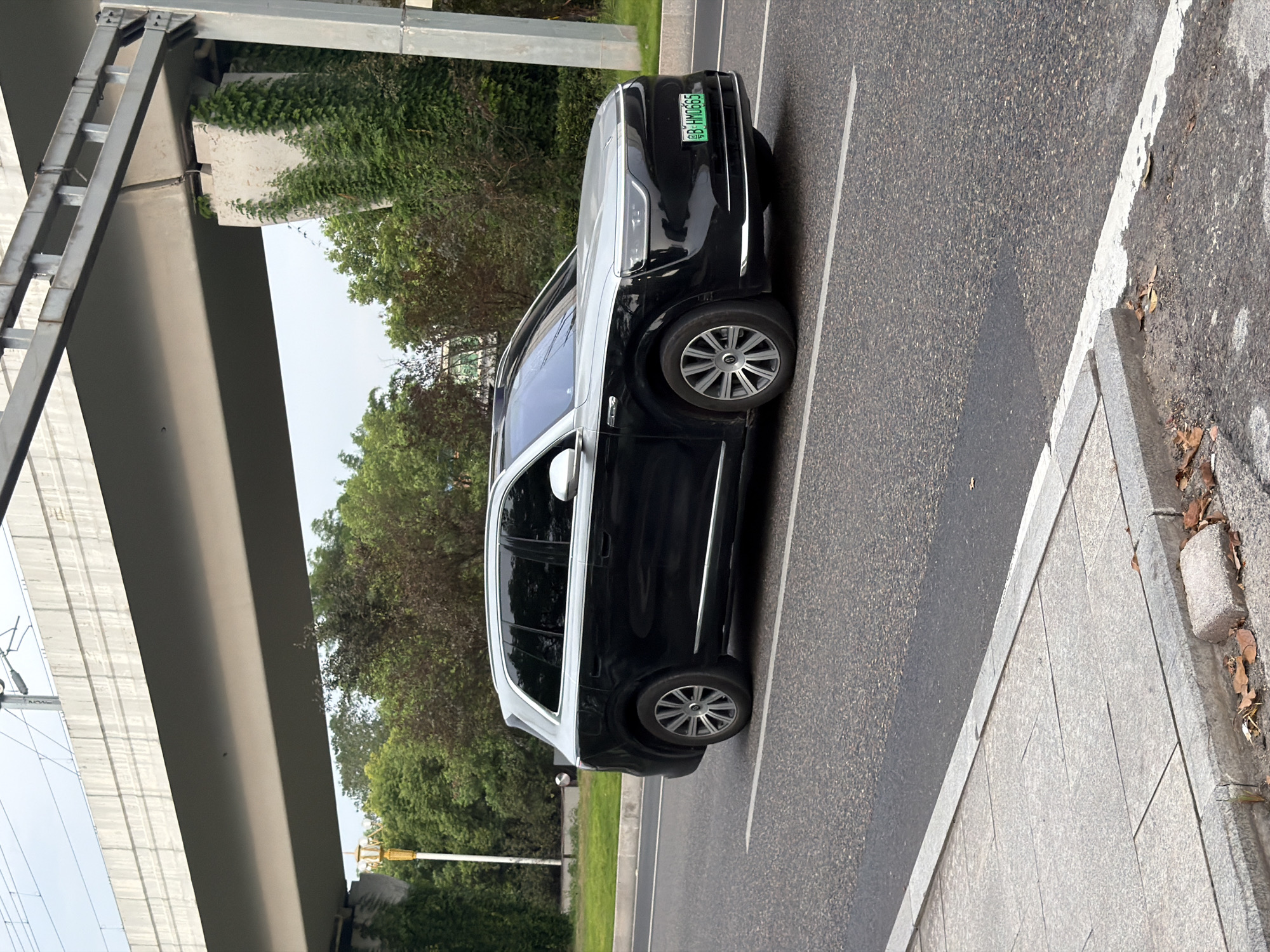 An all-wheel-drive vehicle with a black lower body and a silver upper body is traveling on a paved road. The vehicle has tinted windows and silver rims. A green license plate that reads "B HM0665" is visible on the front of the vehicle. The road is bordered by a sidewalk on the left. In the background, there are trees and an elevated overpass with overhead wires. The scene appears to be in a city. The presence of the license plate format suggests a Chinese origin, and the specific format might be indicative of a particular region or city within China. While the image itself does not contain explicit markers for Jiangjin, China, such a scene is plausible within that urban environment.