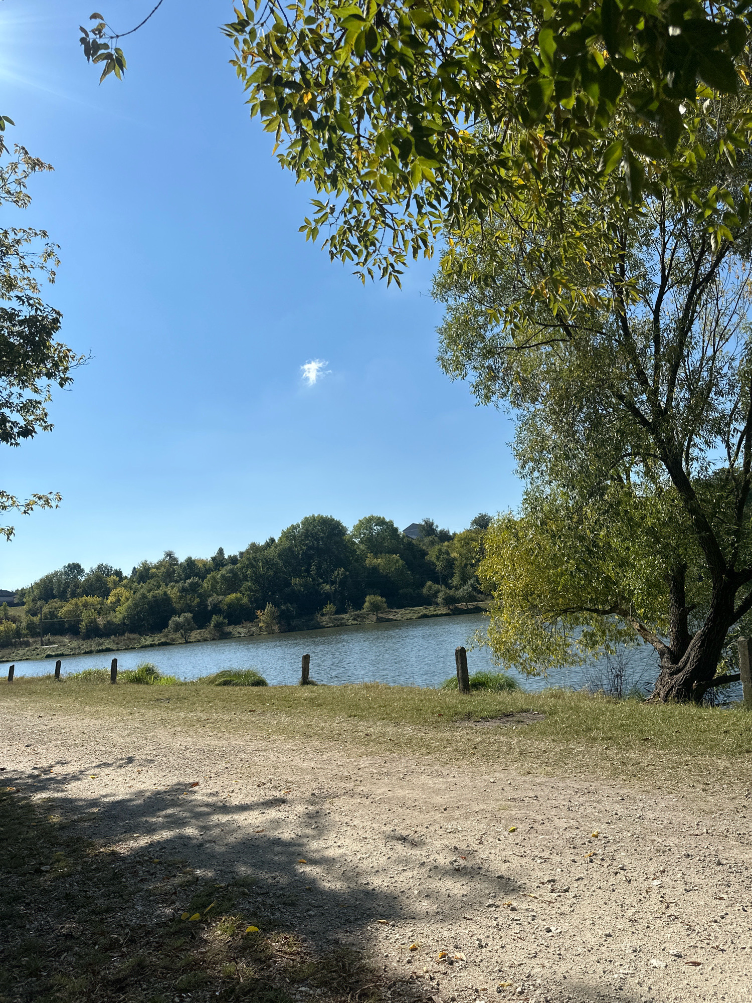 The image depicts a serene natural landscape near Makiv, Ukraine. The scene is dominated by a tranquil body of water, likely a lake or river, reflecting the clear blue sky above. A lush, green treeline borders the opposite bank, creating a picturesque backdrop. In the foreground, a dirt path is visible, casting dappled shadows from overhead foliage. Several wooden posts are strategically placed along the edge of the water, suggesting a boundary or a simple scenic marker. The overall atmosphere is one of peaceful, unspoiled nature.
