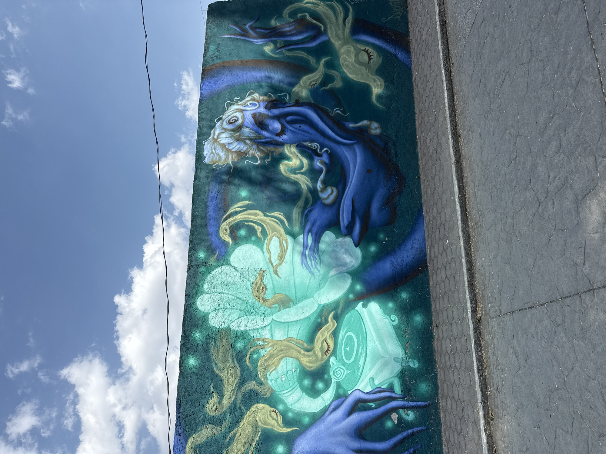 A vertical mural is affixed to the side of a building, presumably in San Andrés Cholula, Mexico. The mural features an otherworldly design in shades of blue, white, and green. A serpentine figure, seemingly composed of flowing fabric, with wispy white hair dominates the upper portion. Below it is a glowing turquoise flower-like form and other ethereal figures with closed eyes and hair that resembles smoke. The mural is on a wall made of small square tiles. The building is adjacent to a rough, grey concrete surface. Above the mural, a section of a blue sky with scattered white clouds is visible. A thin electrical wire is present against the sky and mural.