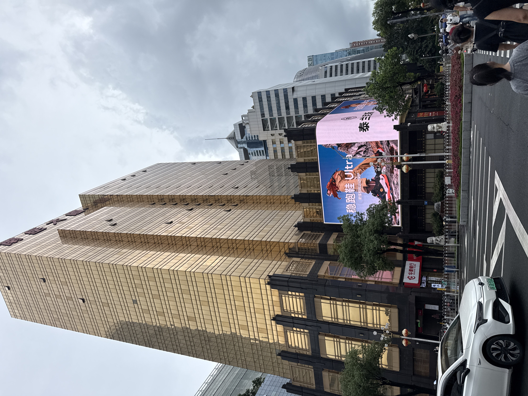 A medium shot captures a street scene in Tianhe District, China, dominated by a tall, golden glass-covered building. A large digital billboard is mounted on the lower section of the building, displaying an advertisement for athletic shoes, featuring a person in an orange jacket and black sneakers. To the right of the billboard, a pink advertisement banner is visible. In front of the building, trees and decorative plants line the sidewalk. A white car is parked on the right side of the street, facing left. Pedestrians are crossing the street in the foreground, with one person wearing a grey t-shirt and another in a black shirt with a backpack. The sky is overcast with clouds.