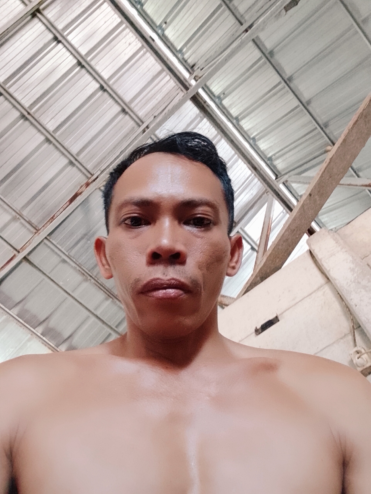 A shirtless man is shown from the chest up in an indoor location. The man has dark hair and skin. He is looking directly at the camera. The ceiling is composed of corrugated metal sheets with wooden beams. The location is in Bukit Ketok, Indonesia.