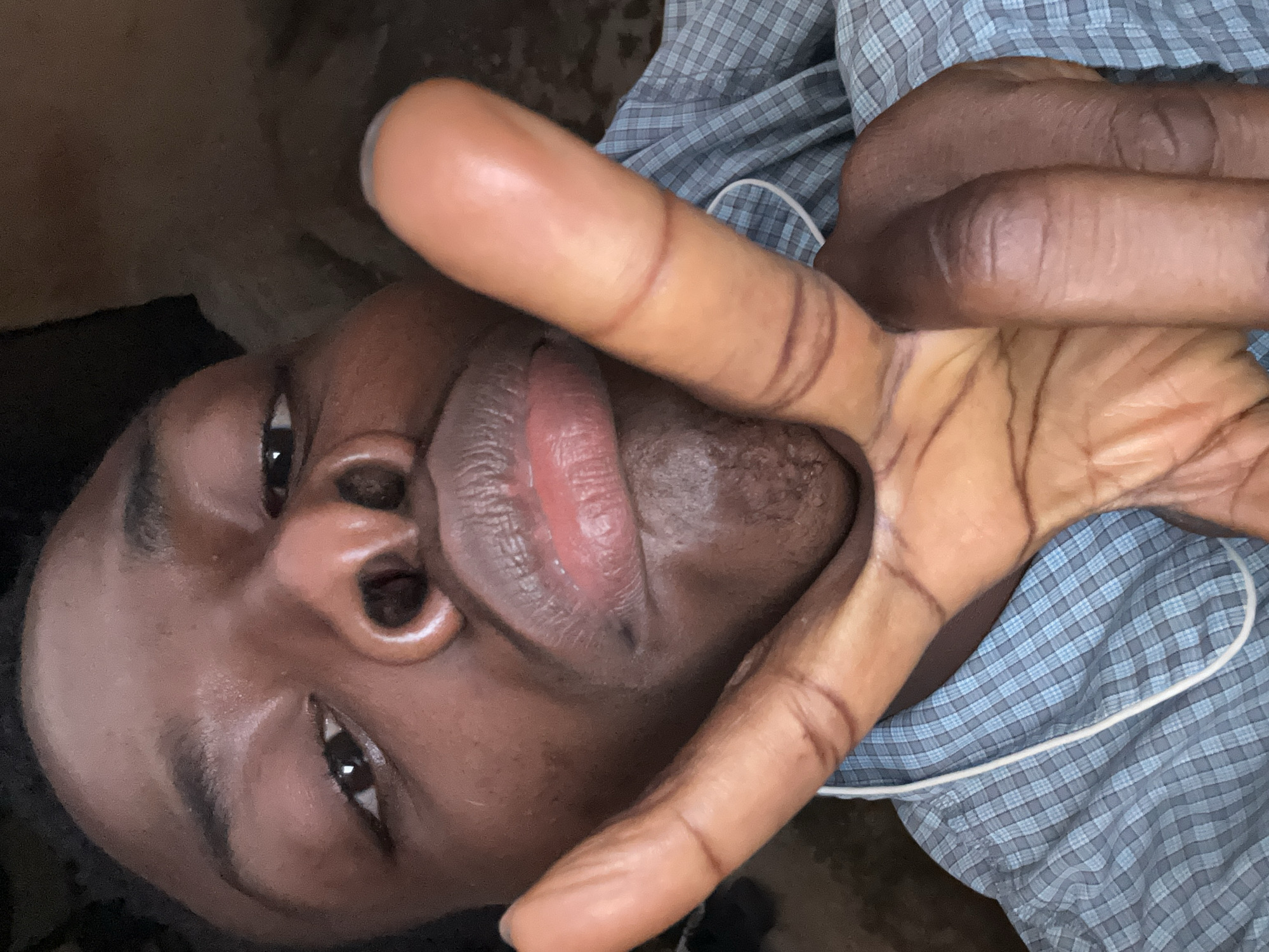 A close-up, indoor photograph shows a dark-skinned individual with their face at an angle. Their right hand is raised, obscuring part of their face with the index and middle fingers extended in a V-shape. The person is wearing a blue and white checkered shirt. No specific location in the city of Kirana, Nigeria, is identifiable.