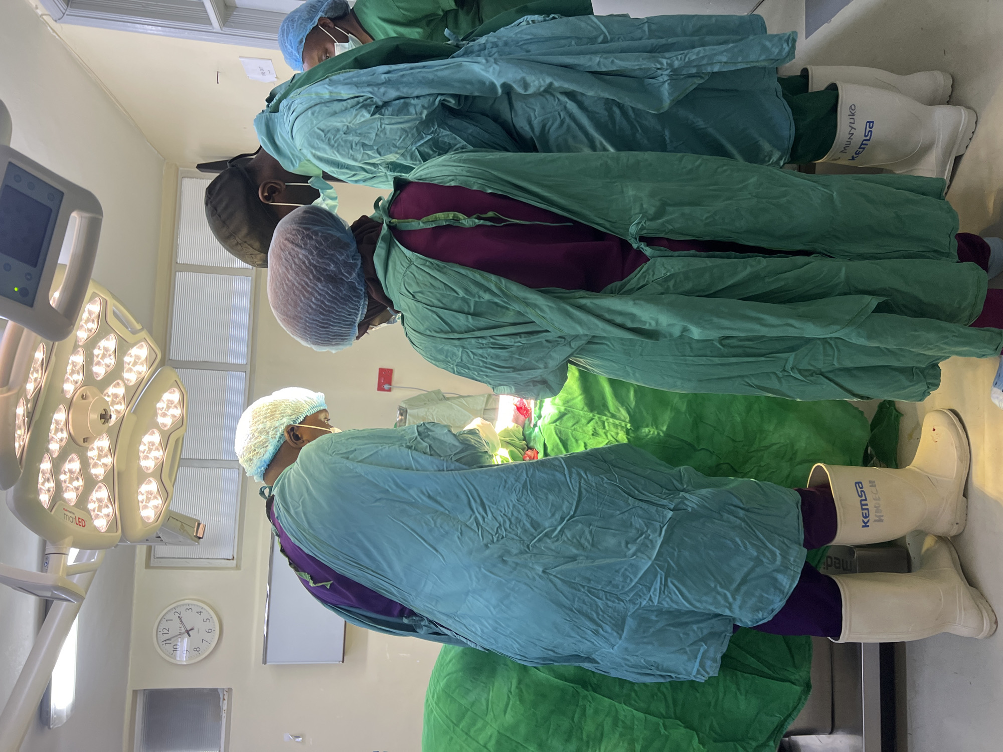 In an operating room, three medical personnel dressed in scrubs, surgical caps, masks, and white boots are performing a surgical procedure. A surgical light shines on the surgical site. There is medical equipment present, including a monitor, and a clock on the wall reads approximately 10:50. The word "Kemsa" is visible on the boots of the person closest to the camera. The location is the city of Nairobi, Kenya, due to "Munyuko kemsa" being written on the boots, which indicates the boots are from the Kenyan supplier.