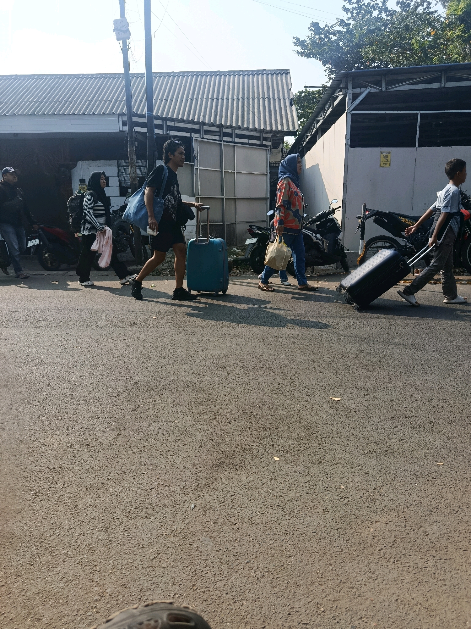 In Jepara, Indonesia, a group of people are walking on a paved road. The scene is outdoors, with a building and trees in the background under a bright sky. A man in a black t-shirt and shorts is pulling a large, turquoise suitcase with wheels. Beside him, a woman wearing a blue headscarf and a colorful patterned shirt is carrying a beige tote bag. To the left, a woman in a striped shirt and black pants, also with a headscarf, is walking and carrying a pink item. A man in a black jacket and jeans is visible further to the left, standing near motorcycles. Another person, a young boy in a light grey shirt, is walking on the right, pulling a black suitcase. The road surface is asphalt, with some scattered leaves. The building in the background has a corrugated metal roof.