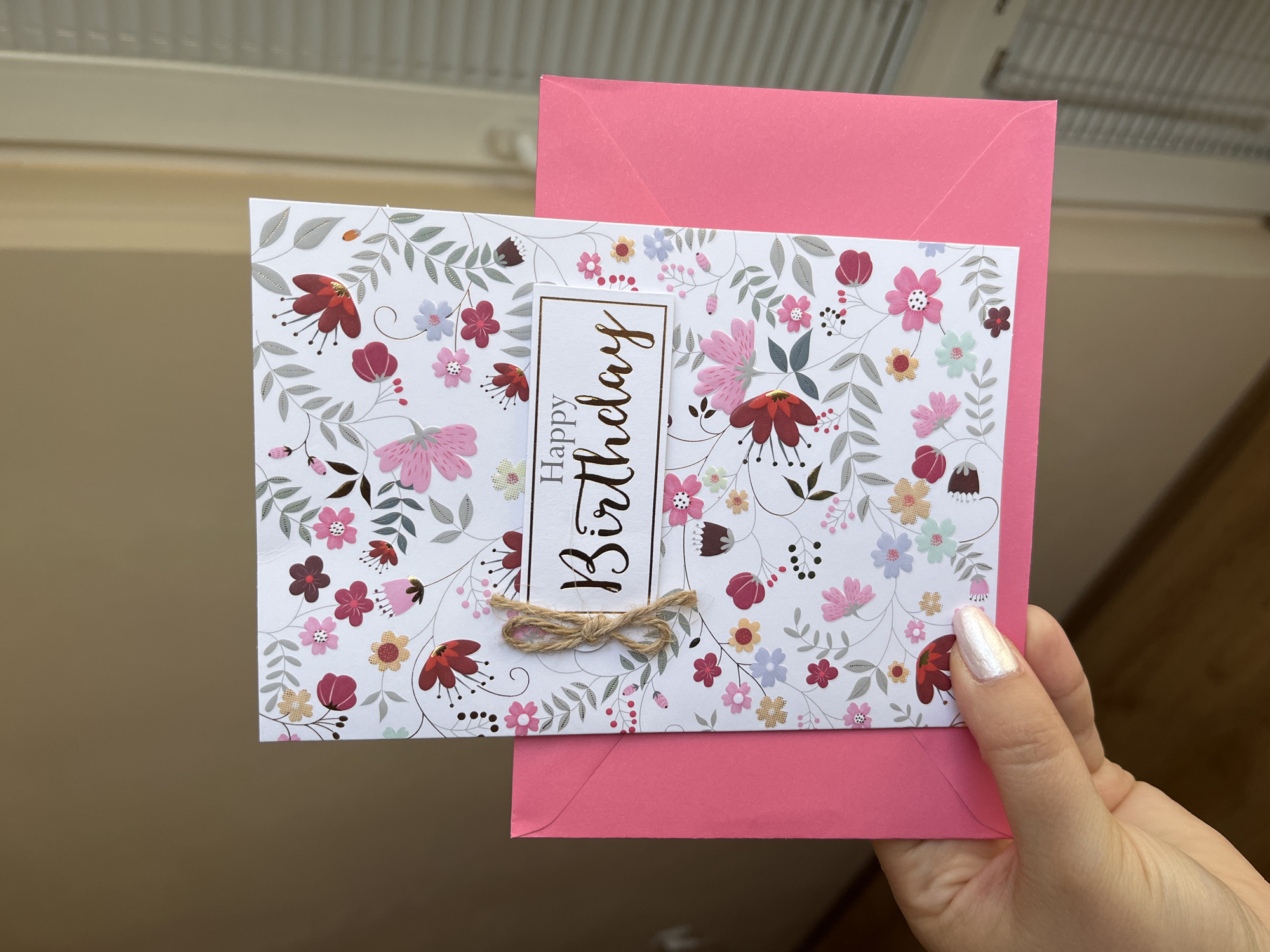 A hand with white nail polish holds a floral birthday card over a pink envelope. The card has a white background with a delicate pattern of pink, red, and blue flowers and green leaves. The words "Happy Birthday" are printed in gold cursive lettering on a white rectangle in the center of the card, tied with twine. The pink envelope is visible beneath the card, with its pointed flap extending upwards. The background is a plain, light brown wall. There is no identifiable location within the city of Plovdiv, Bulgaria.