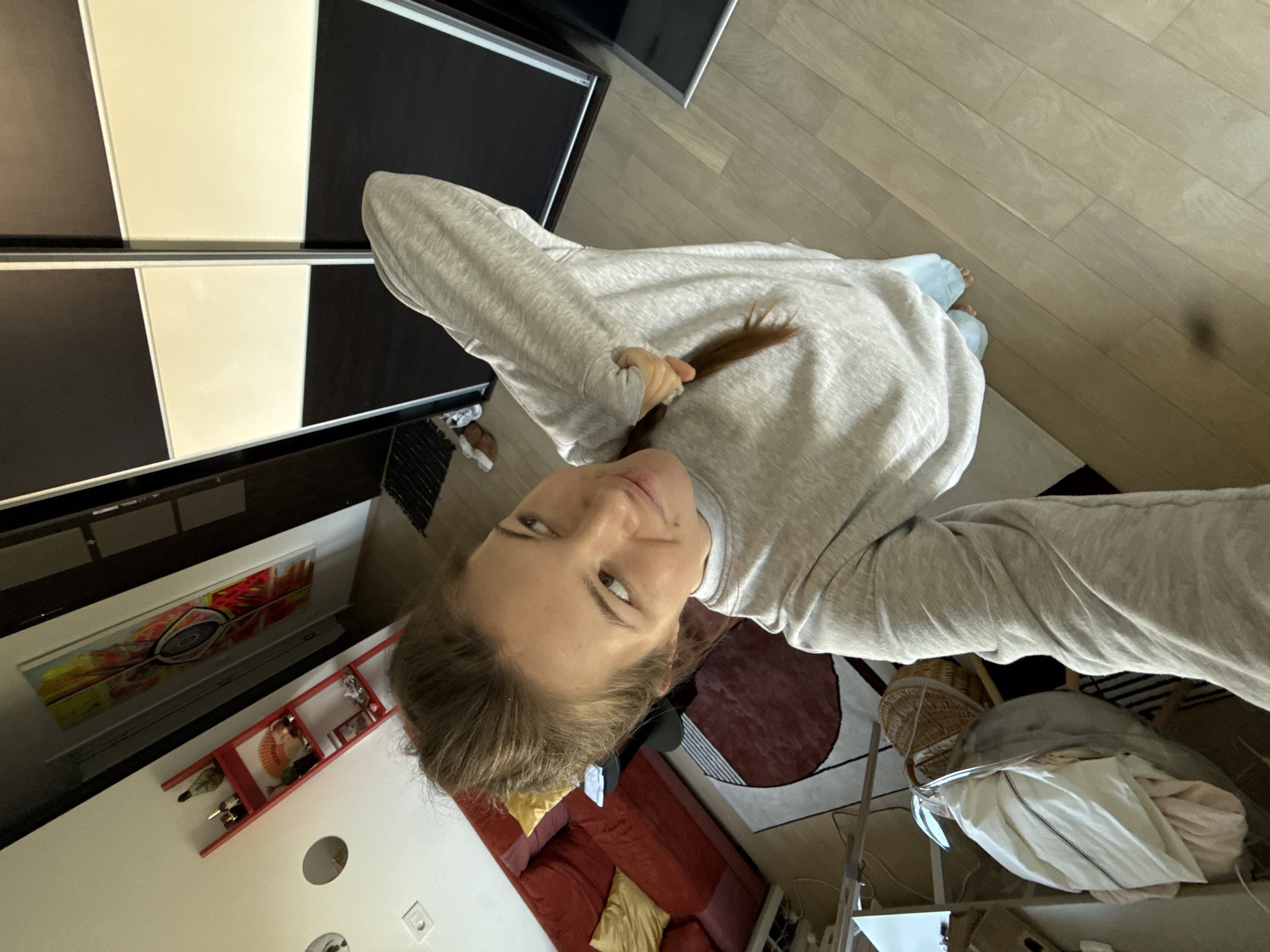 A high-angle selfie of a young adult female with light brown hair wearing a light gray long-sleeved sweatshirt and light blue jeans. The flooring is light-colored wood. Behind her is a room containing a red sofa with yellow pillows, a red wall-mounted shelf containing wine bottles, and a photograph on the wall. A black and white wardrobe closet is to the left, and part of a patterned rug is also visible. The location is likely an apartment within the Vozdovac Urban Municipality, Serbia.