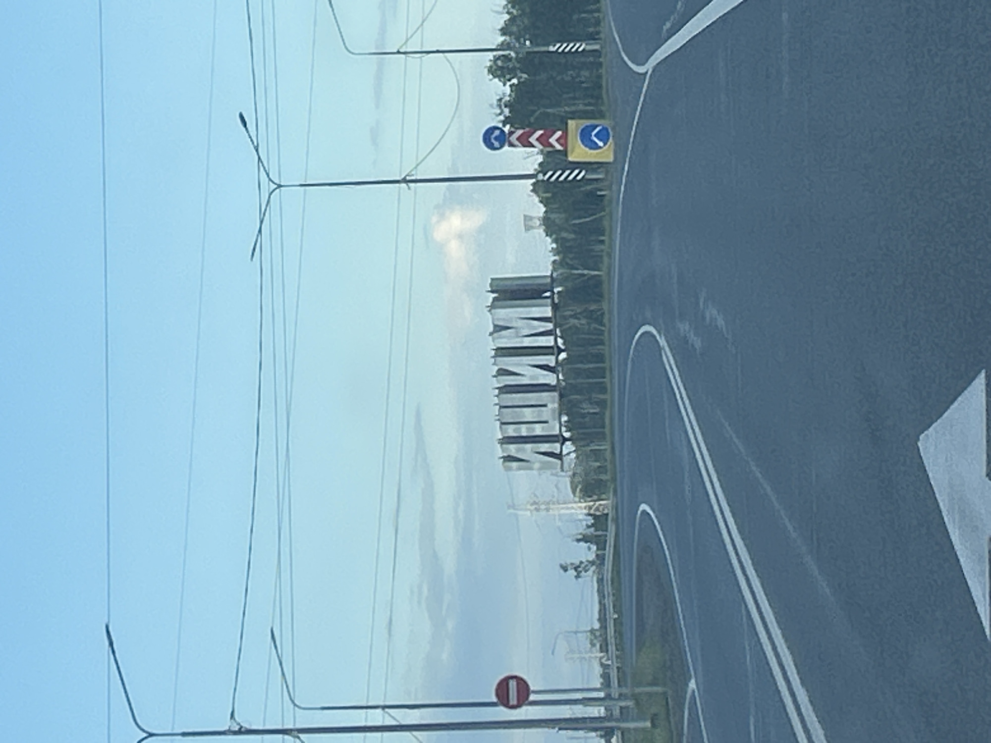 This is a photograph taken outdoors, likely during the daytime given the bright sky. The image captures a section of a paved road with white lane markings, suggesting it is a highway or a similar main thoroughfare. The road curves to the right, indicating a bend or an intersection. On the left side of the road, there are several tall streetlights and overhead power lines, suggesting an urban or suburban setting. In the distance, a large, modern building with a distinctive architectural design is visible, appearing to be made of many stacked horizontal elements.
Among the notable details are traffic signs. One sign shows a series of red and white chevron arrows pointing to the right, indicating a sharp turn or a need to move to the right. Adjacent to this sign is a yellow square sign with a blue arrow pointing to the right, likely a directional sign. Further down, a circular red and white sign is visible, which typically signifies a prohibition, such as "Do Not Enter" or "No Entry." The sky is mostly clear with some scattered clouds, and the overall lighting suggests it is not dawn or dusk. The image is oriented vertically, as if the camera was held sideways. The background includes trees and foliage, hinting at a natural landscape bordering the road. No people are visible in the frame. The activity depicted is simply the scene of a road with associated infrastructure and signage, suggesting a location where traffic management is in place. There is no visible text other than the symbols on the traffic signs. The location context is given as Lozovoe, Russia.