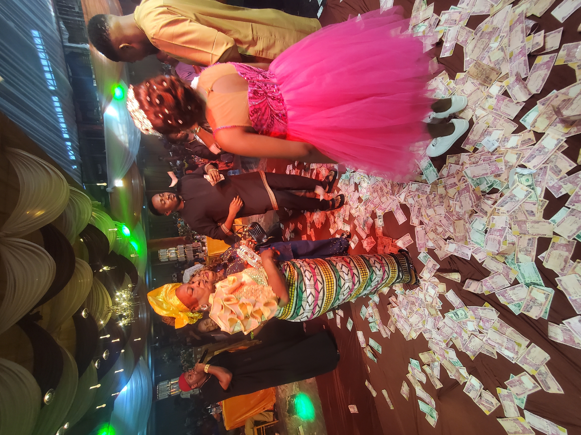 The image captures a celebratory scene within a venue, possibly located in Unknown, Unknown. Scattered across the floor, which is covered with a brown material, are numerous bank notes. Several individuals are present in the scene. One woman, adorned with a yellow headwrap and a vibrant, patterned dress, lies on the money-covered floor. Another woman, wearing a bright pink dress, stands near her. A man in a dark suit is also lying on the floor, while other people are standing and engaging in the celebration. The venue is decorated with black, white, and yellow drapery, and a chandelier hangs from the ceiling. A man in a red cap is singing into a microphone, suggesting a live performance. The lighting in the venue is varied, with green lights visible in the background.