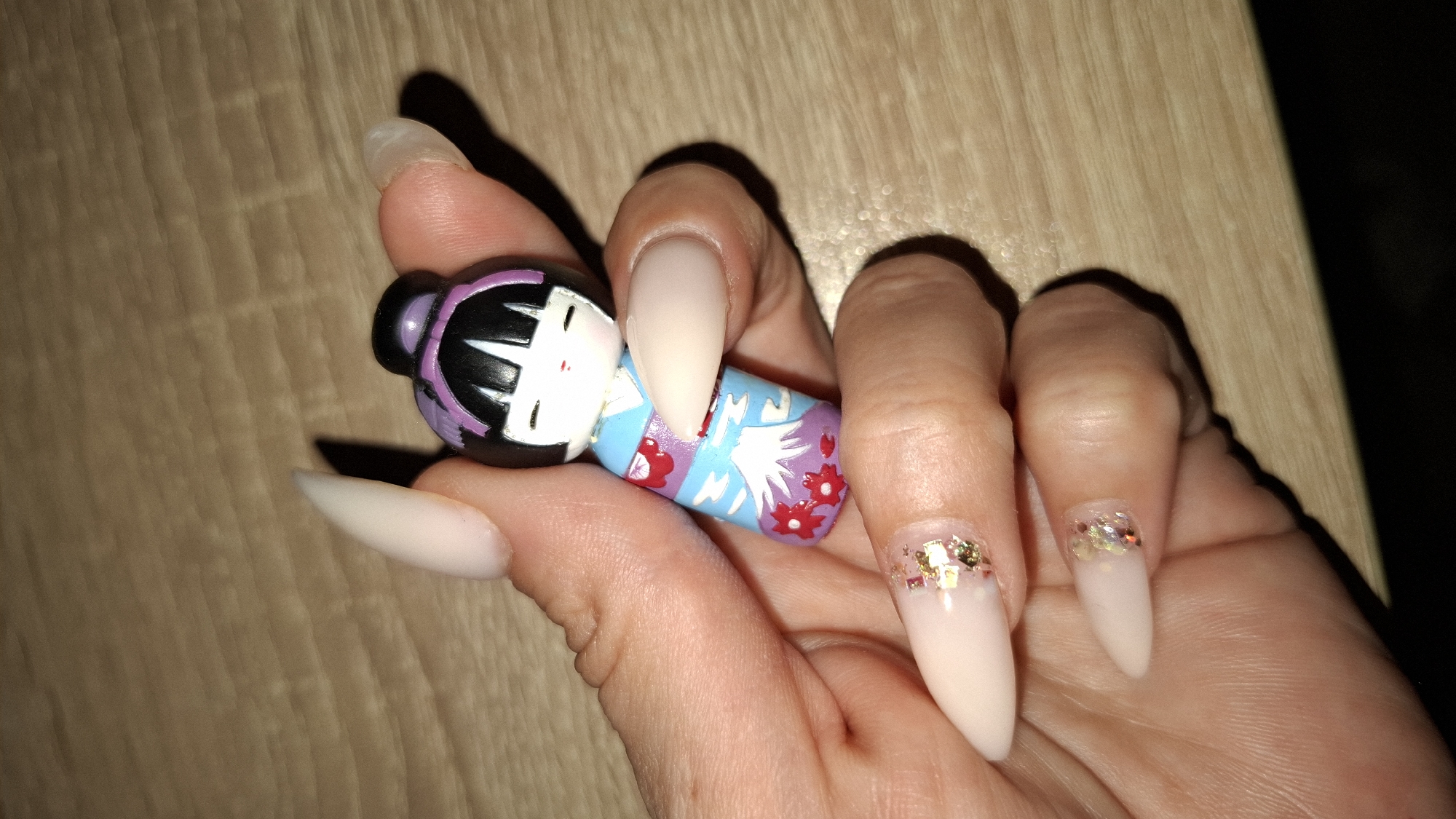 A person's hand is holding a small, painted Kokeshi doll on a wooden surface. The person has long, almond-shaped fingernails, two of which are decorated with gold glitter near the base. The doll is painted with a traditional Japanese design, featuring a black head with bangs, a white face, and a blue kimono adorned with red flowers. The hand is angled, with the fingers curled around the doll. The wooden surface is light brown with a visible grain pattern. The lighting in the scene appears dim, casting shadows around the hand and doll. There is no indication of the location being Sofia, Bulgaria.
