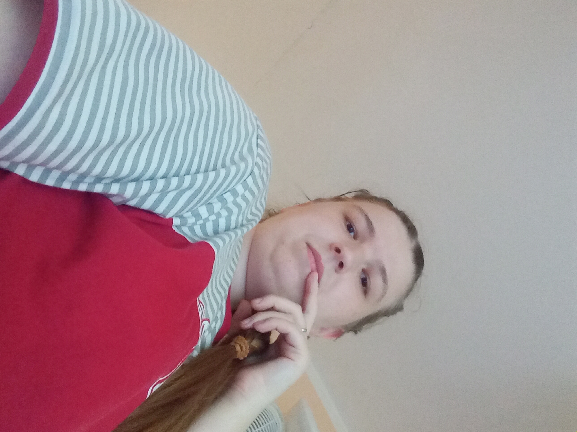 The image shows a low-angle, close-up shot of a young person wearing a red shirt with grey and white striped sleeves. The person has a pigtail and is resting their chin on their hand, with their finger touching their lips. Their eyes are looking towards the camera, and they have a thoughtful or contemplative expression. The background is a plain, light-colored wall. The lighting appears to be indoor, with no specific indicators of time of day or weather. There is no visible text in the image. The overall impression is of a casual, personal photograph.